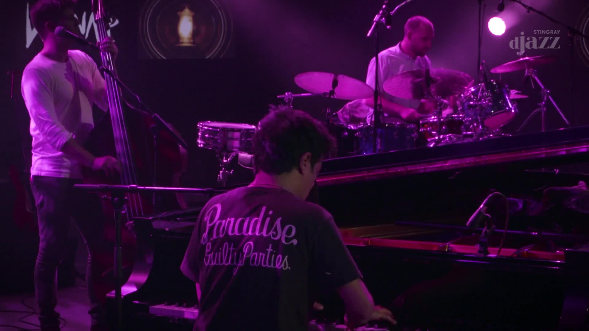 The bassist is leaning into his instrument, fingers moving across the strings. A pianist, wearing a shirt with "Paradise Guilty Parties" on the back, plays the keys of a grand piano. The drummer is a blur of motion behind his kit. The bassist is leaning into his instrument, fingers moving across the strings. A pianist, wearing a shirt with "Paradise Guilty Parties" on the back, plays the keys of a grand piano. The drummer is a blur of motion behind his kit.
