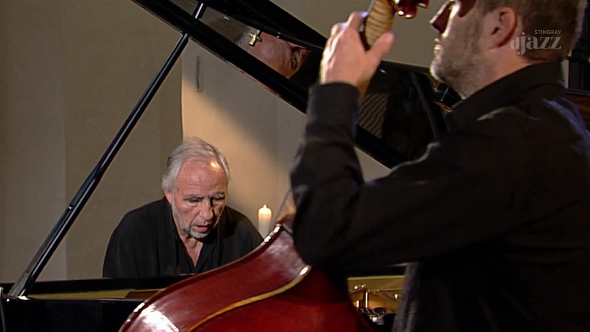 A pianist with grey hair plays a grand piano while a bassist, his back to the camera, bows his upright bass. The Stingray DJAZZ logo is visible in the upper right corner. A pianist with grey hair plays a grand piano while a bassist, his back to the camera, bows his upright bass. The Stingray DJAZZ logo is visible in the upper right corner.