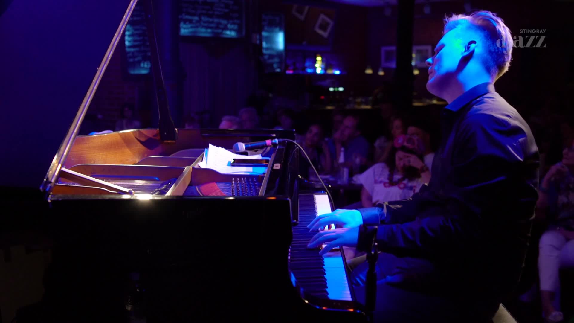 A pianist's hands dance across the keys of a grand piano, bathed in the cool blue light of a Canadian jazz club. The audience, a collection of faces in the dim background, watches the performance, likely a part of the Stingray DJAZZ broadcast. A pianist's hands dance across the keys of a grand piano, bathed in the cool blue light of a Canadian jazz club. The audience, a collection of faces in the dim background, watches the performance, likely a part of the Stingray DJAZZ broadcast.