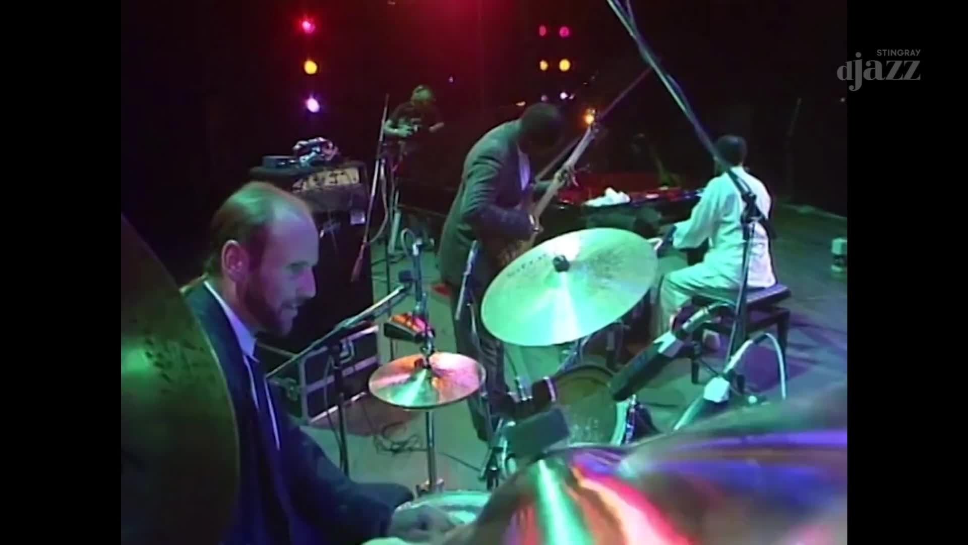 The drummer, a man with a beard, taps his cymbals while a bassist in a suit plays his instrument. A keyboardist in a white robe sits at his station, bathed in stage lights. The drummer, a man with a beard, taps his cymbals while a bassist in a suit plays his instrument. A keyboardist in a white robe sits at his station, bathed in stage lights.
