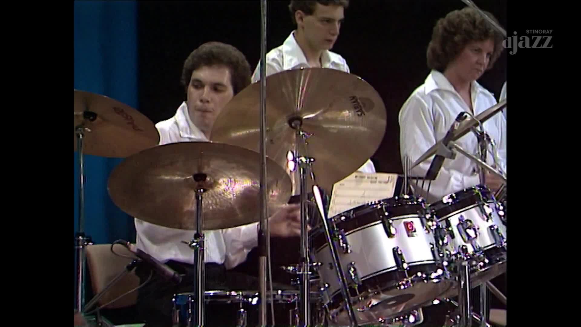 The drummer's sticks are a blur as he hits the cymbals, his focus intense. Beside him, a woman in a white shirt leans into her drums, the Stingray Jazz logo visible on the right. The drummer's sticks are a blur as he hits the cymbals, his focus intense. Beside him, a woman in a white shirt leans into her drums, the Stingray Jazz logo visible on the right.