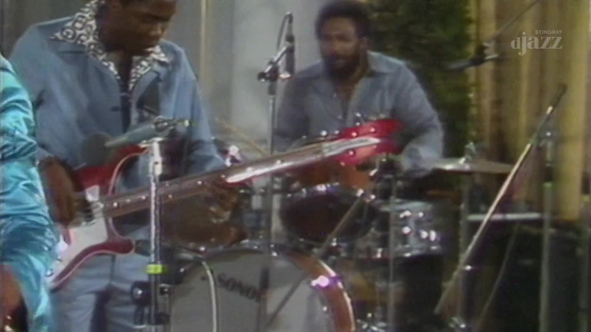 A bassist in a blue jacket plays a red guitar, his fingers moving across the fretboard. Behind him, a drummer with a beard keeps time on his kit, the cymbals catching the light. A bassist in a blue jacket plays a red guitar, his fingers moving across the fretboard. Behind him, a drummer with a beard keeps time on his kit, the cymbals catching the light.