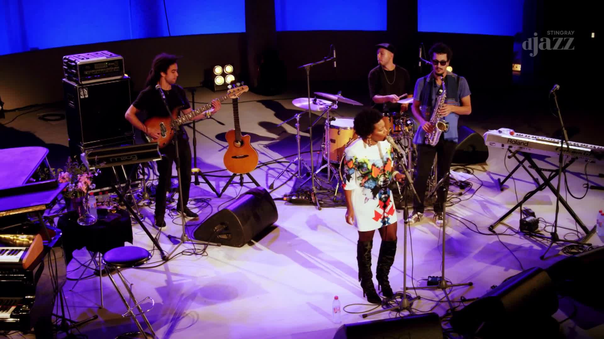 A singer in a vibrant dress stands center stage, microphone in hand, while a saxophonist plays beside her. A guitarist on the left strums his instrument, and a drummer is visible behind them, all bathed in the stage lights of this Stingray DJAZZ broadcast from Canada. A singer in a vibrant dress stands center stage, microphone in hand, while a saxophonist plays beside her. A guitarist on the left strums his instrument, and a drummer is visible behind them, all bathed in the stage lights of this Stingray DJAZZ broadcast from Canada.