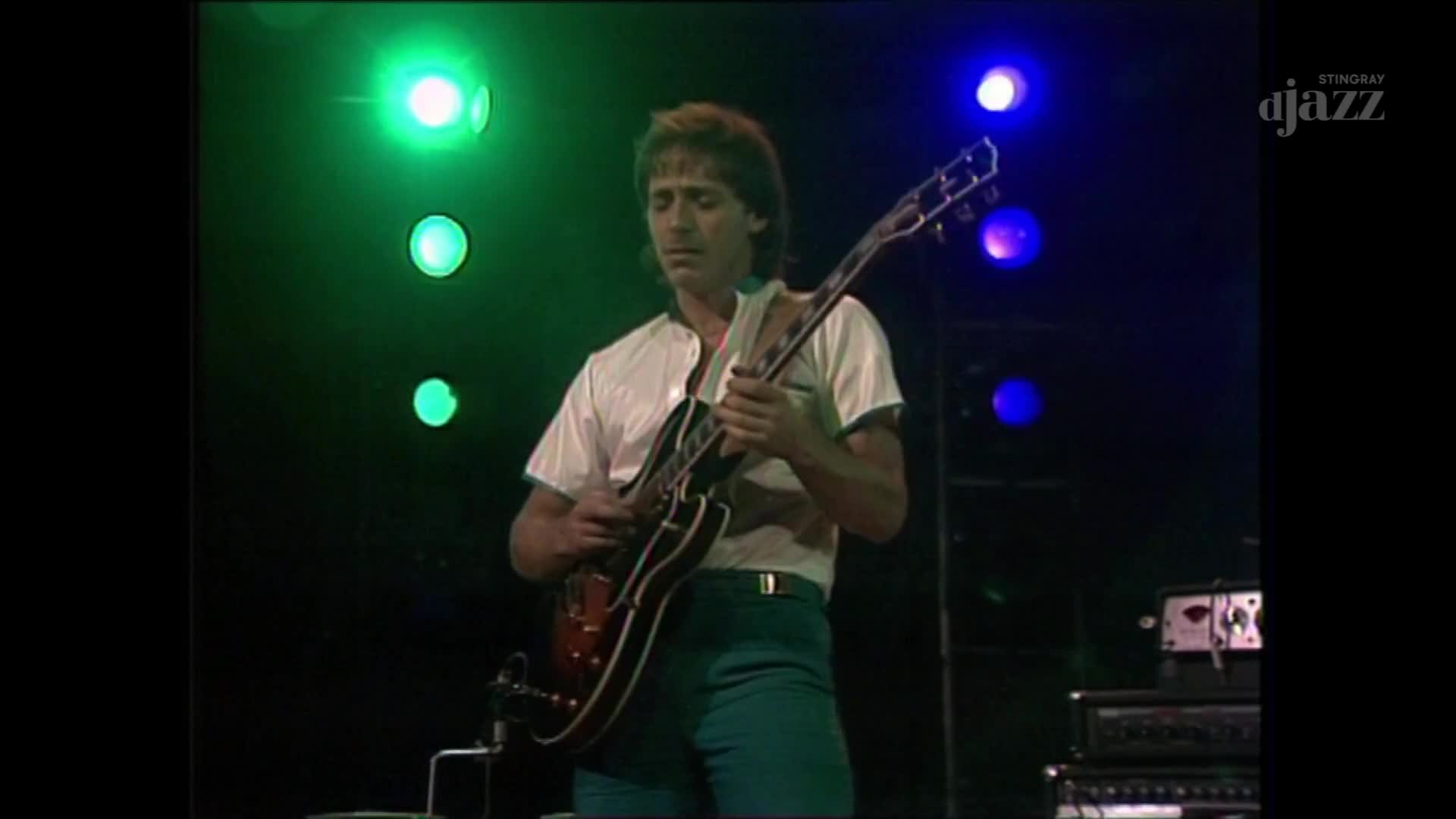 A guitarist stands center stage, his fingers dancing on the fretboard of a dark, hollow-body electric guitar. Green and blue lights pulse behind him, illuminating the performance for Stingray DJAZZ.