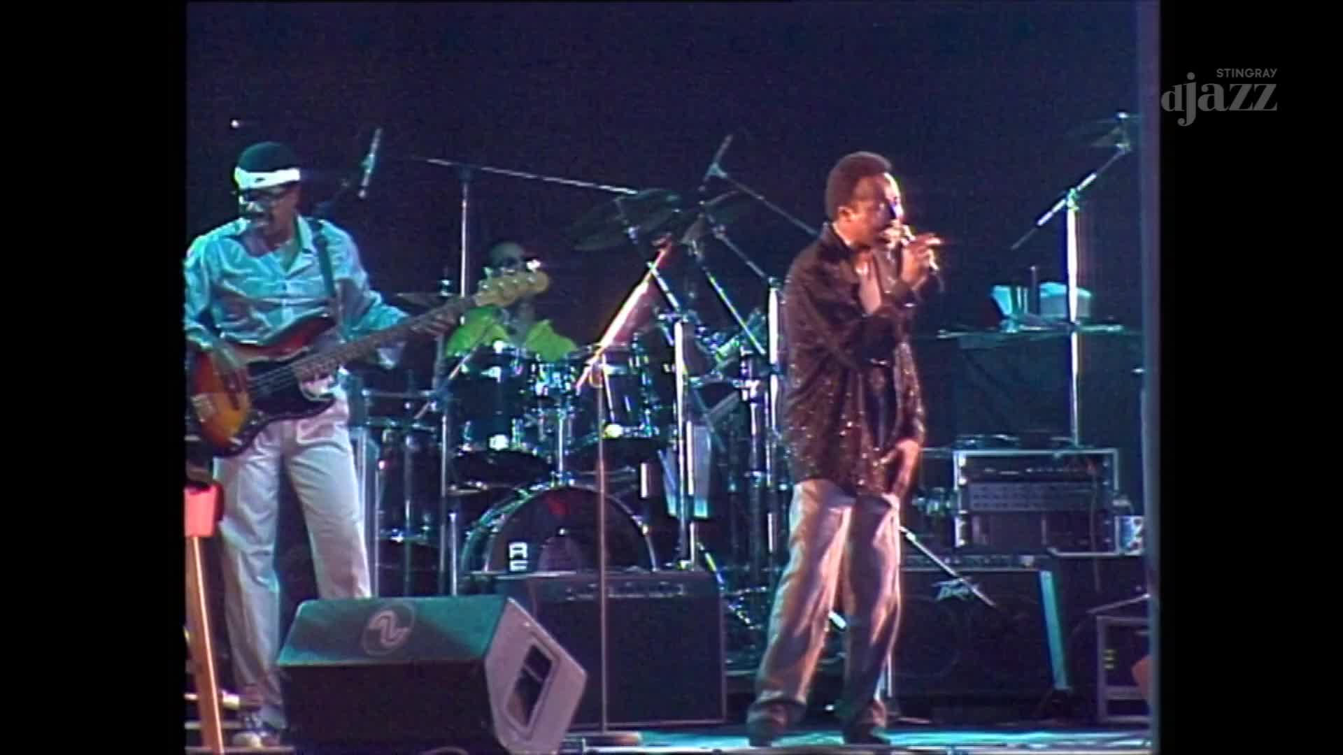 A singer in a sparkling jacket belts into a microphone while a bassist in a light blue shirt plays his instrument. The drummer keeps the beat behind them, all bathed in stage lights.