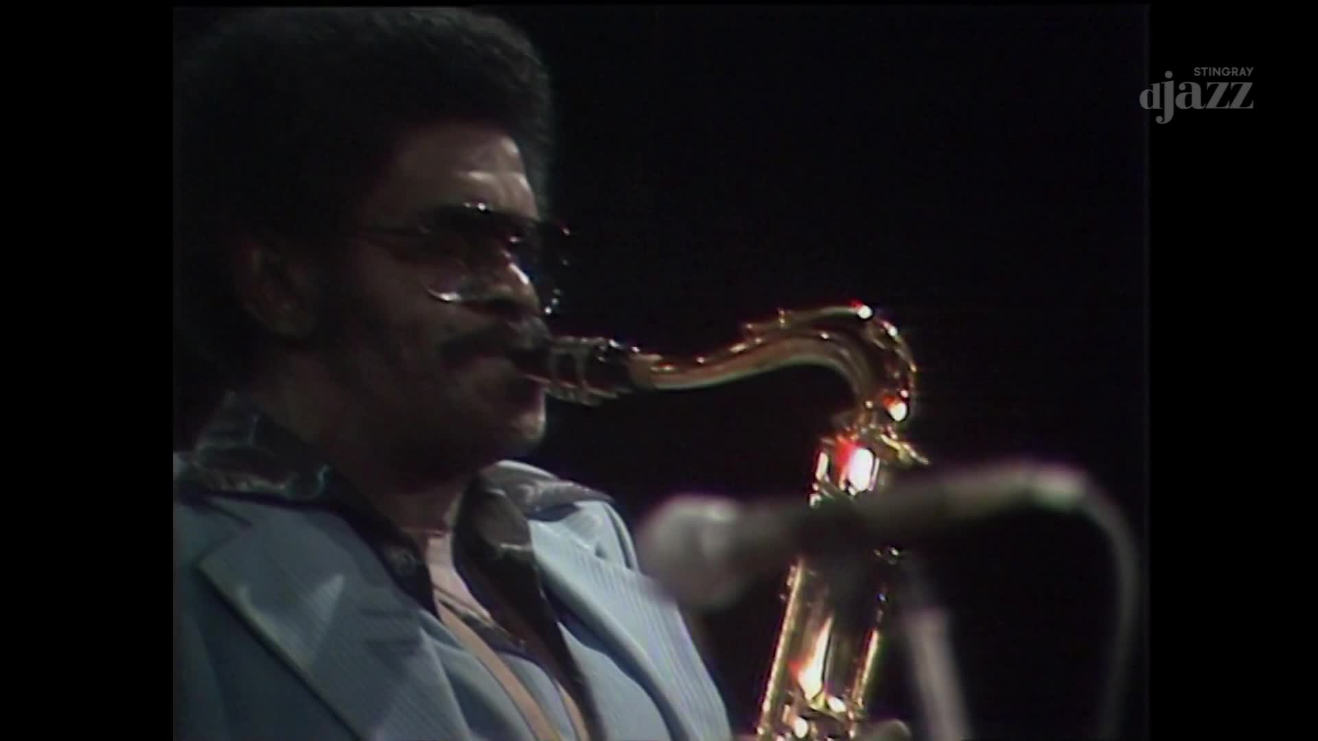A man in a light blue suit plays a saxophone, his fingers moving nimbly on the keys. The brass instrument gleams under the stage lights as he blows into the mouthpiece, filling the air with music.