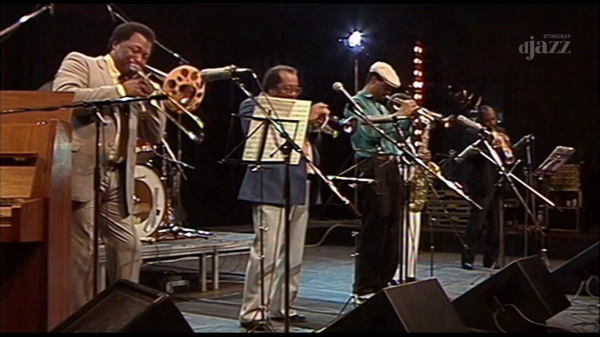 A trombonist in a light suit plays his instrument while a pianist sits nearby. A trumpet player in a green shirt and cap stands next to a saxophonist, all on a stage with bright lights.