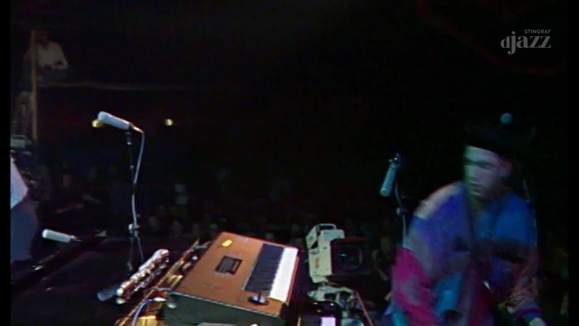 A man in a colorful shirt and hat moves across the stage, his face a blur. Microphones stand ready on stands, and a keyboard sits on a stand, waiting for the Stingray DJAZZ performance to continue.