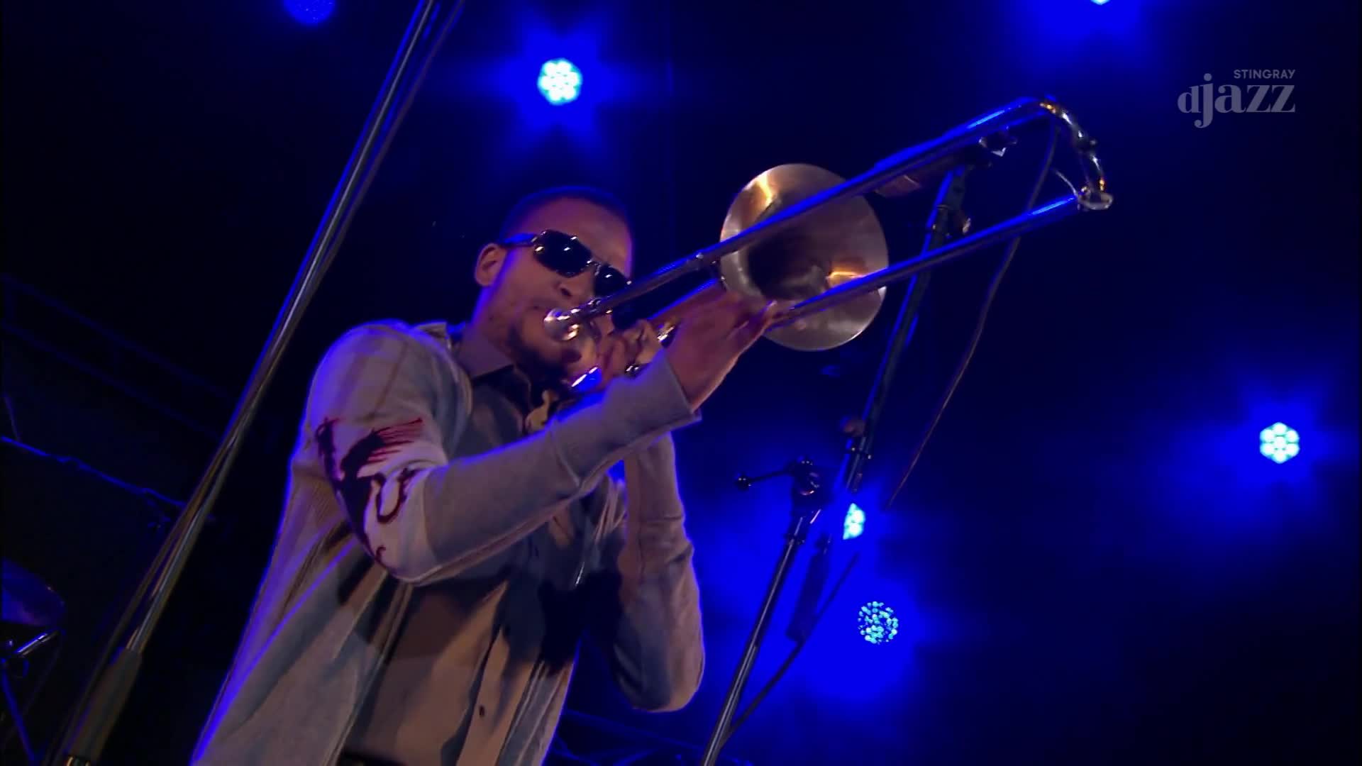 A trombonist, wearing sunglasses, plays his instrument under a wash of blue stage lights. The Stingray DJAZZ logo is visible in the upper right corner of the dark background.