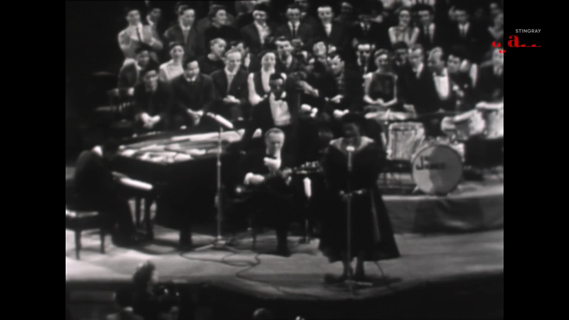 A woman in a dark dress stands at a microphone, her voice filling the hall. A pianist plays a grand piano to her left, while a drummer keeps time behind them.