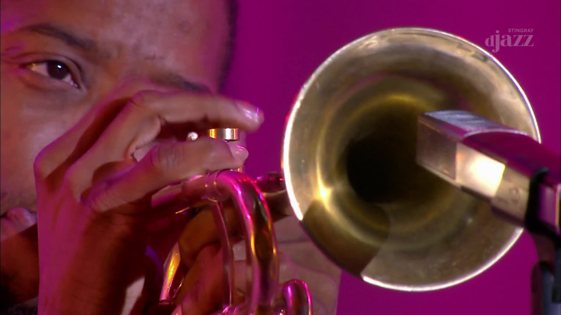 Fingers press down on the valves of a brass trumpet, its bell aimed at a microphone. The musician's focused gaze is visible just above the instrument.