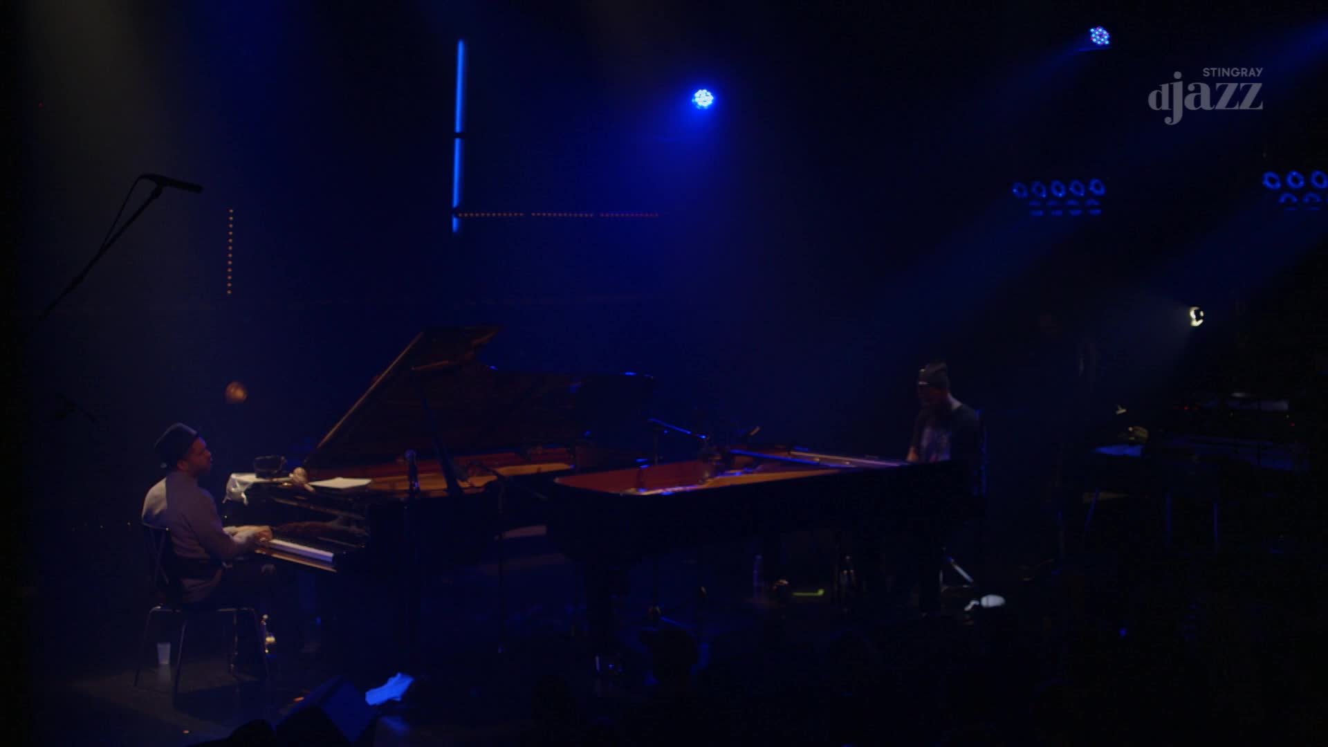 Two pianists are performing on a dimly lit stage, bathed in blue light. The Stingray DJAZZ logo hangs in the upper right corner.