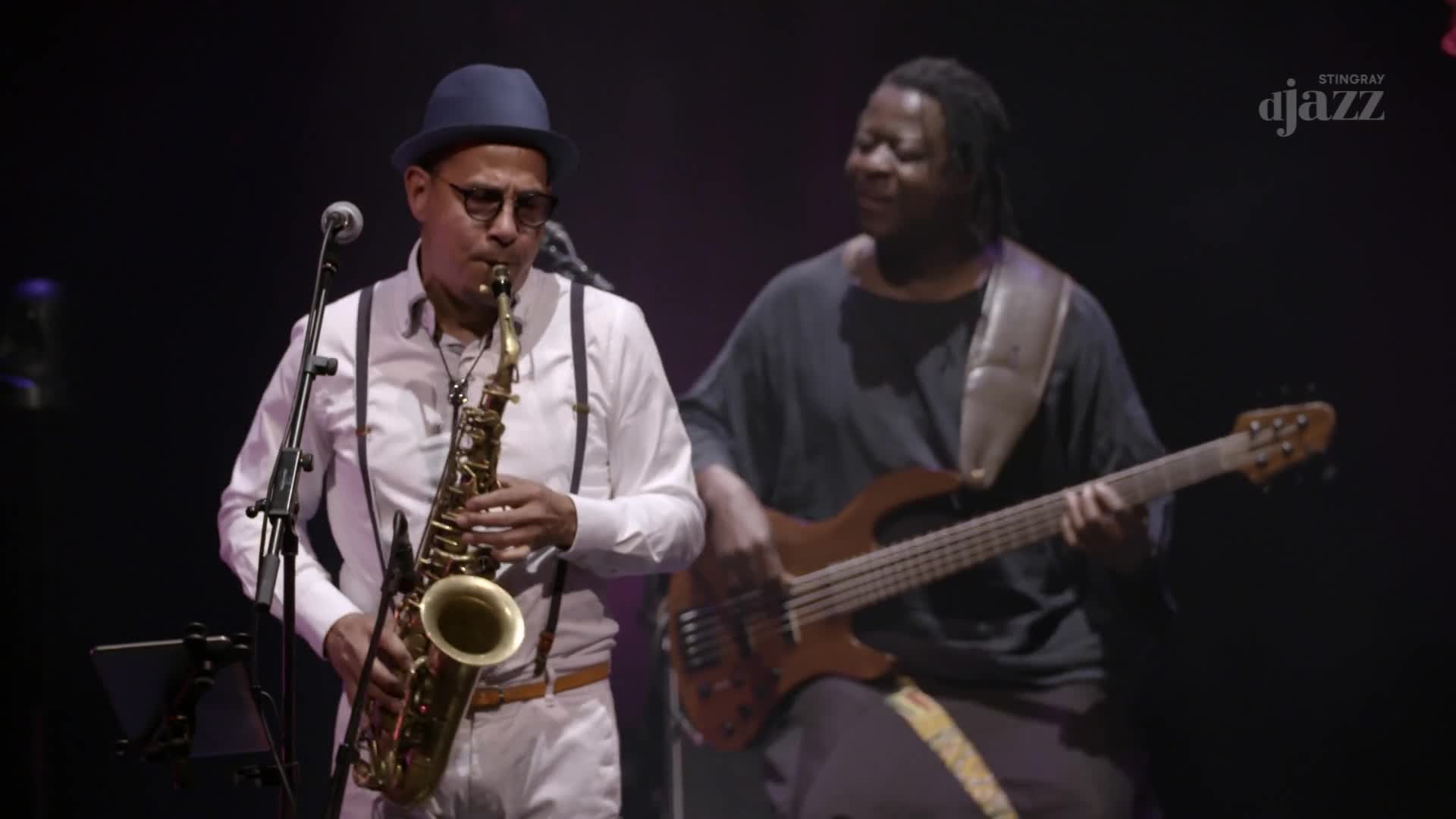A man in a blue hat and suspenders plays a saxophone, his fingers moving nimbly over the keys. Beside him, another musician plays a bass guitar, his head tilted back as he sings along. The Stingray DJAZZ logo is visible in the corner, hinting at a Canadian broadcast.