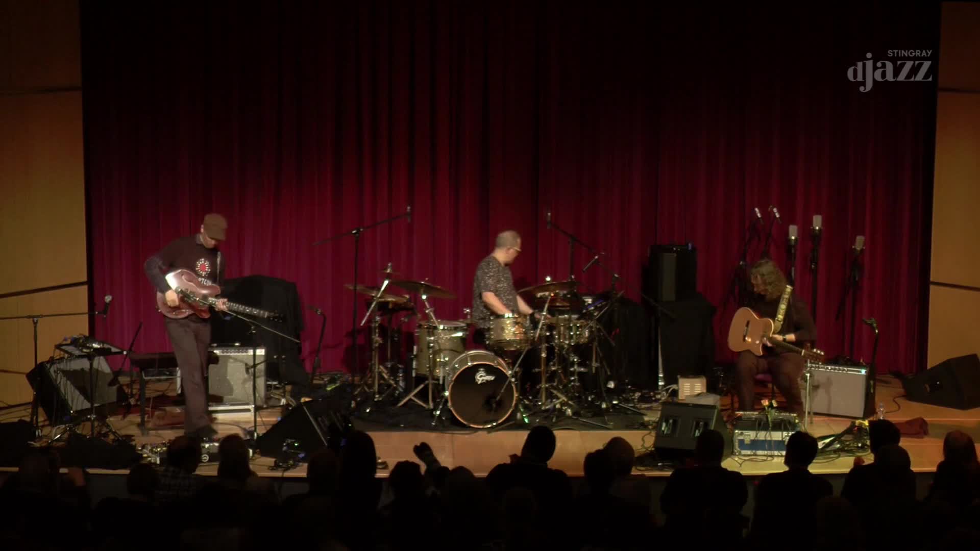 A guitarist stands and plays his instrument, his body swaying slightly to the rhythm. Behind him, a drummer taps out a beat on his kit, while another guitarist sits and strums his guitar. This Canadian jazz trio is performing on a stage with a deep red curtain backdrop, broadcast by Stingray DJAZZ.