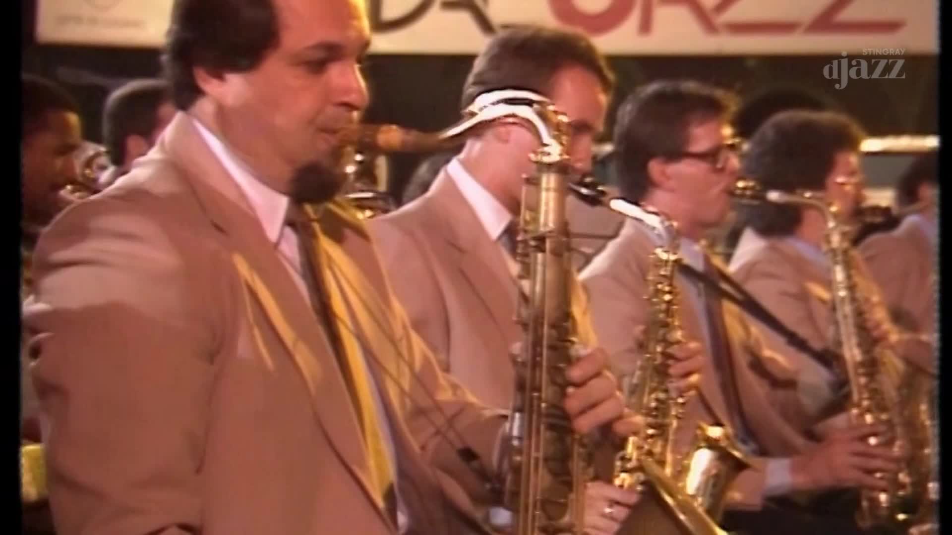 A band plays saxophones on a stage in Canada, the bright lights catching the brass instruments. The musicians, all in matching tan suits, focus on their music for the Stingray DJAZZ broadcast.