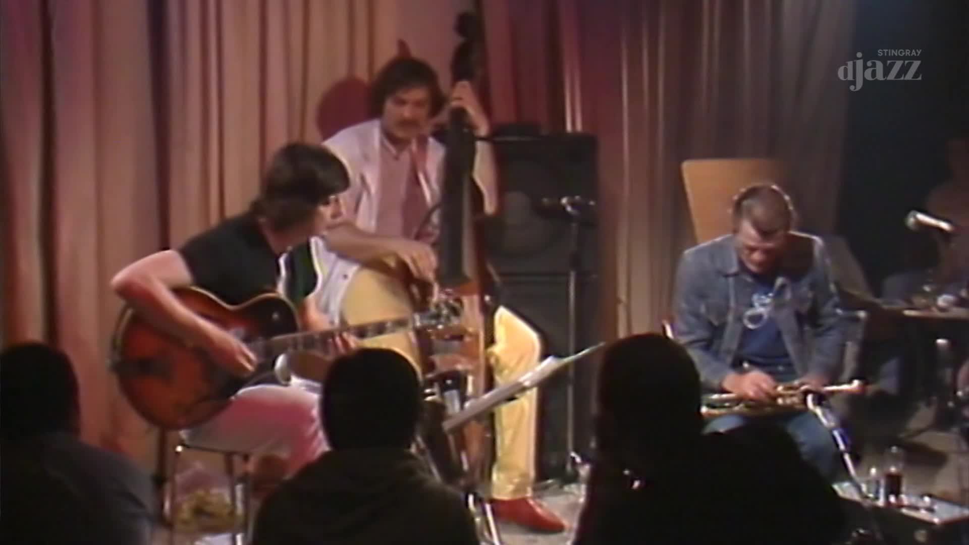 A guitarist in a black t-shirt plays his instrument, while a bassist in bright yellow pants stands behind him. To the right, a man in a denim jacket holds a trumpet.