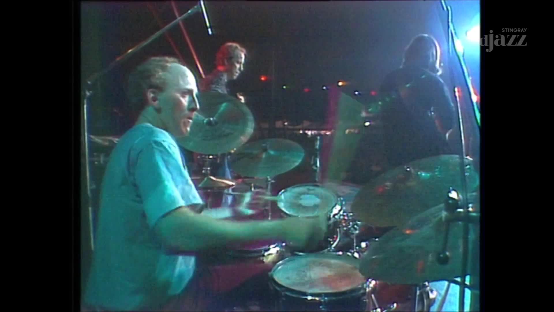 The drummer's hands are a blur as he hits the cymbals, his face focused on the rhythm. Behind him, two other musicians are silhouetted against the stage lights, playing their instruments for Stingray DJAZZ.