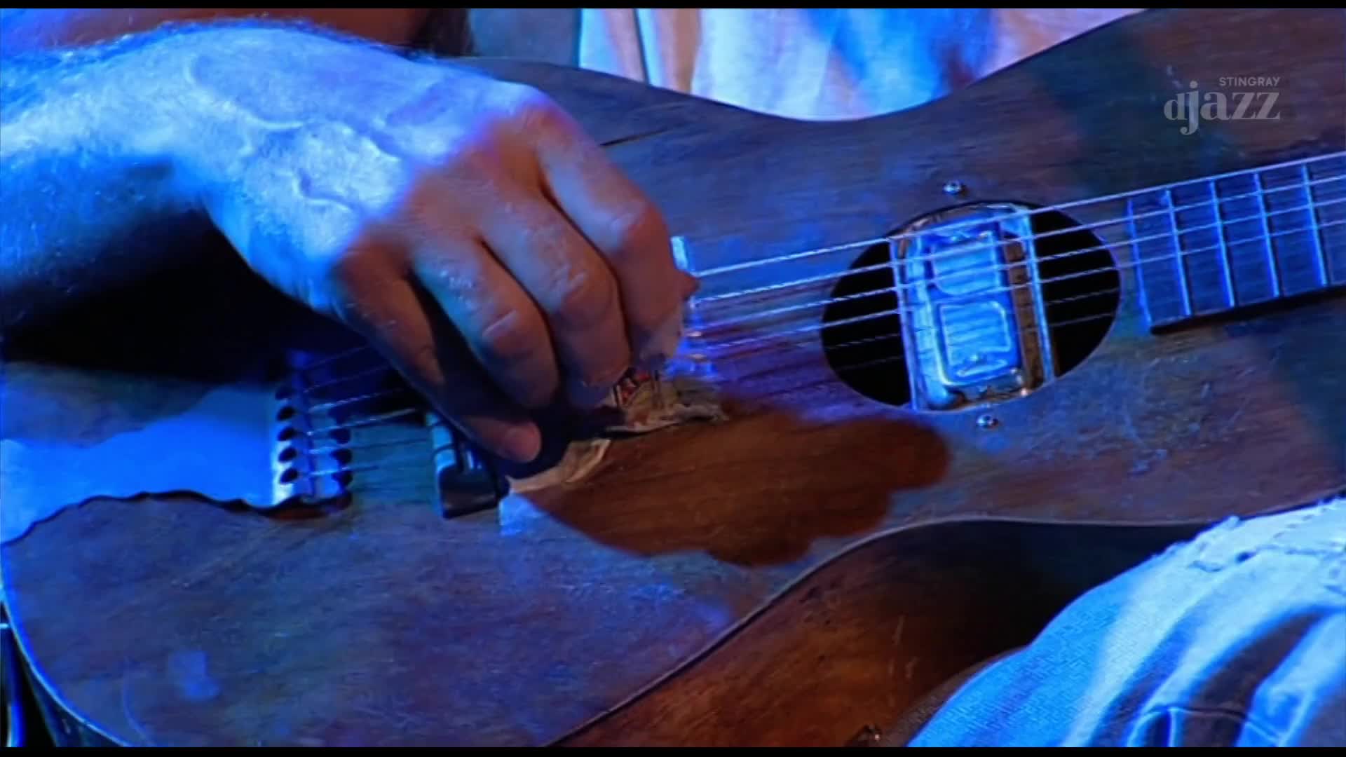 A hand strums an electric guitar, its polished wood gleaming under blue stage lights. The Stingray DJAZZ logo is visible on the instrument.