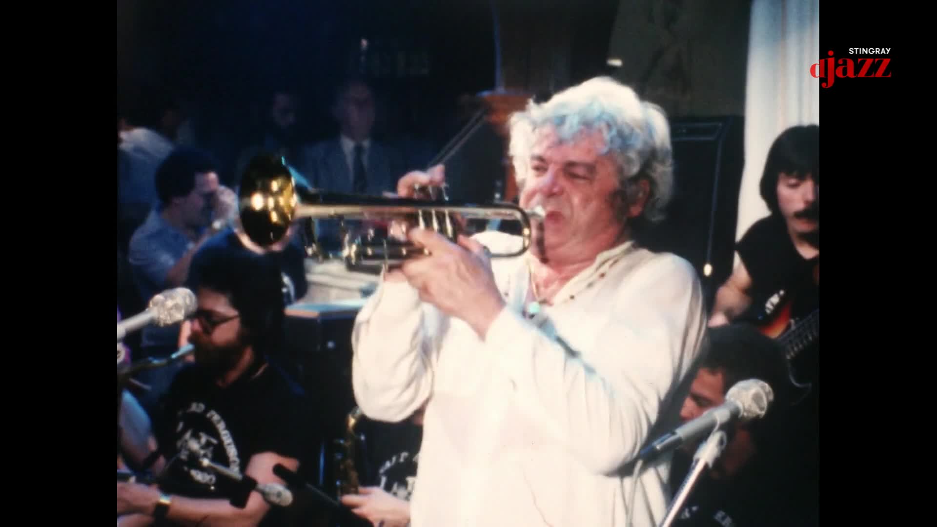 A man with white hair plays a trumpet, his cheeks puffed out. Behind him, a guitarist with a mustache strums his instrument.