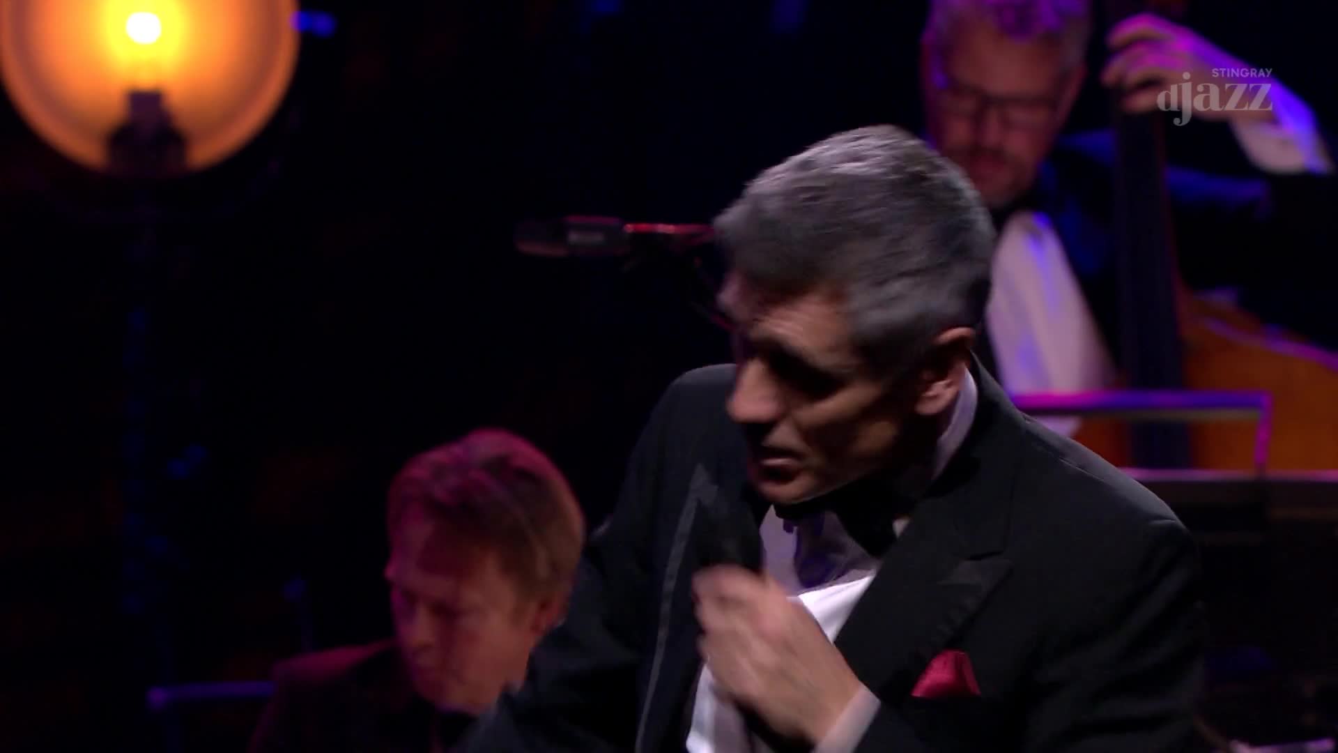 The man in the tuxedo leans forward, his hand adjusting his tie. Behind him, a double bass player stands, his instrument catching the stage light.