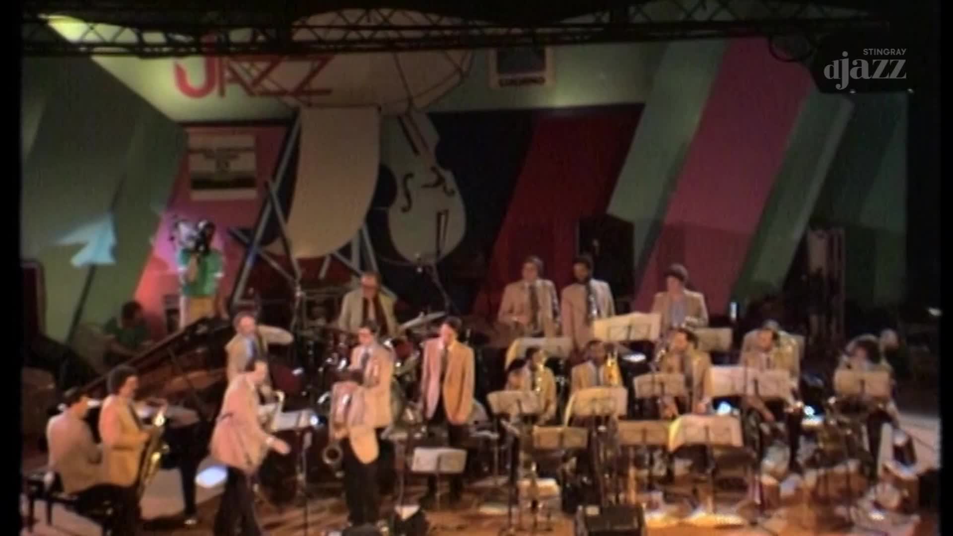A jazz band is playing on stage in front of a colorful backdrop featuring the word "JAZZ." A cameraman in a green shirt stands near the stage's edge, filming the performance.
