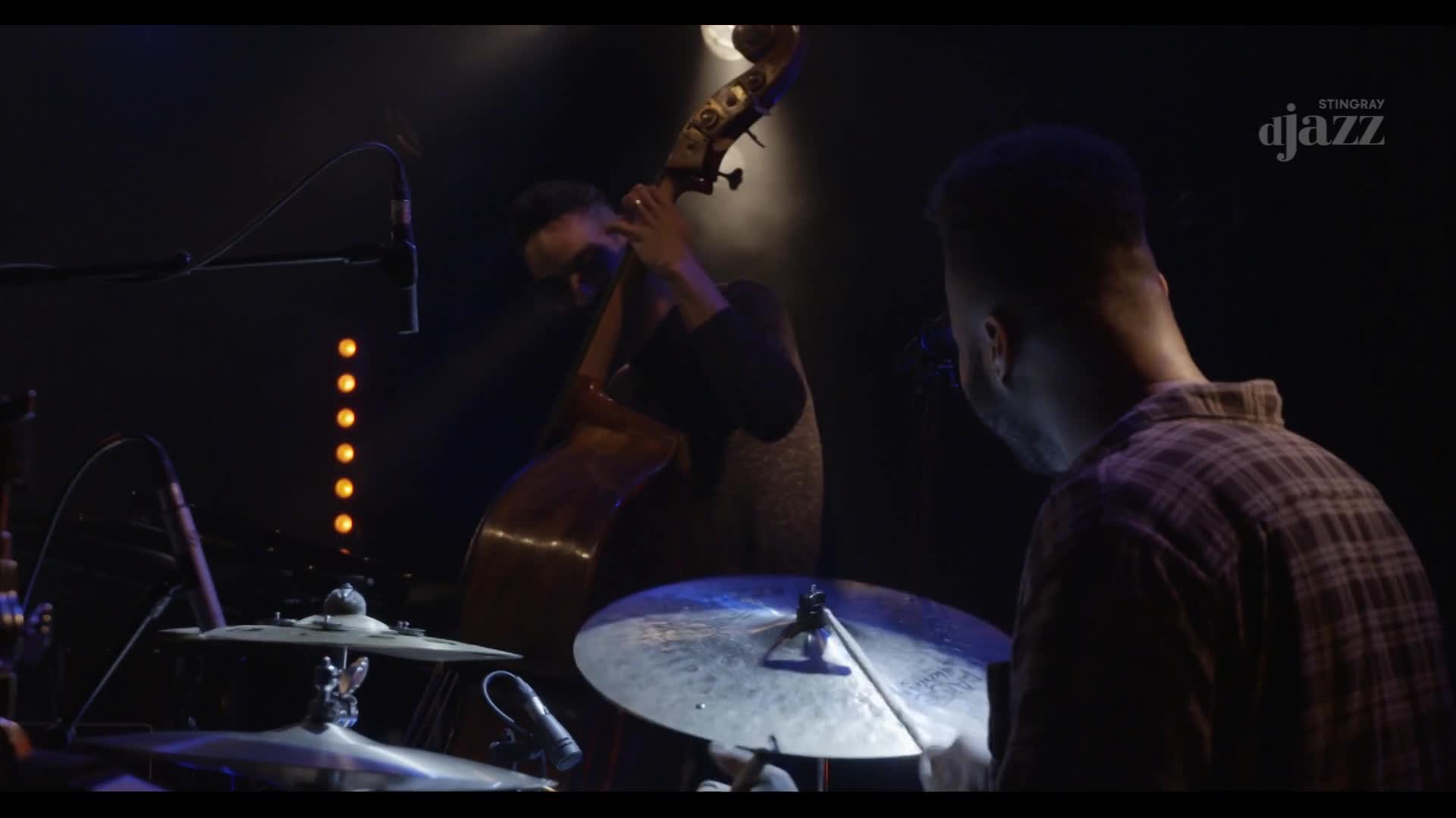A double bass player leans into his instrument under the stage lights. The drummer's stick hovers over a cymbal, ready to strike.