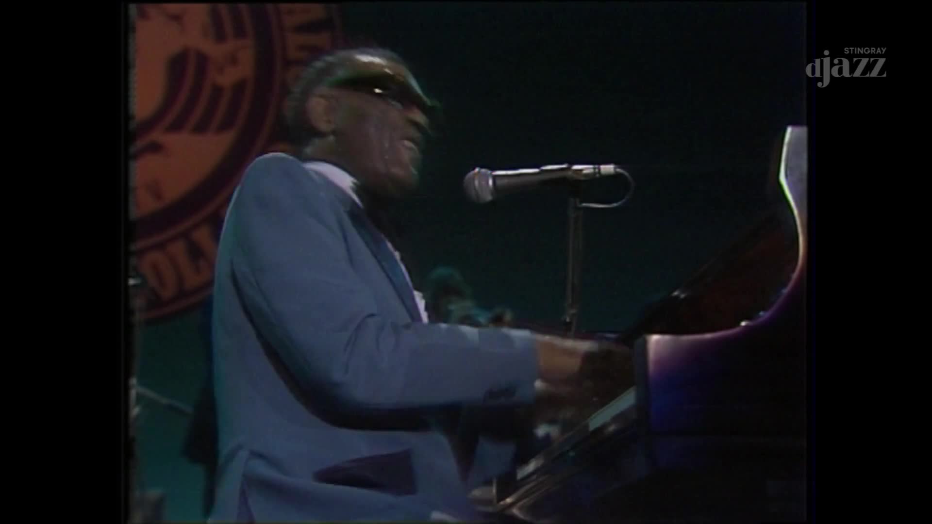 The pianist's hands move across the keys, his sunglasses reflecting the stage lights. A microphone stands ready, positioned just above him.