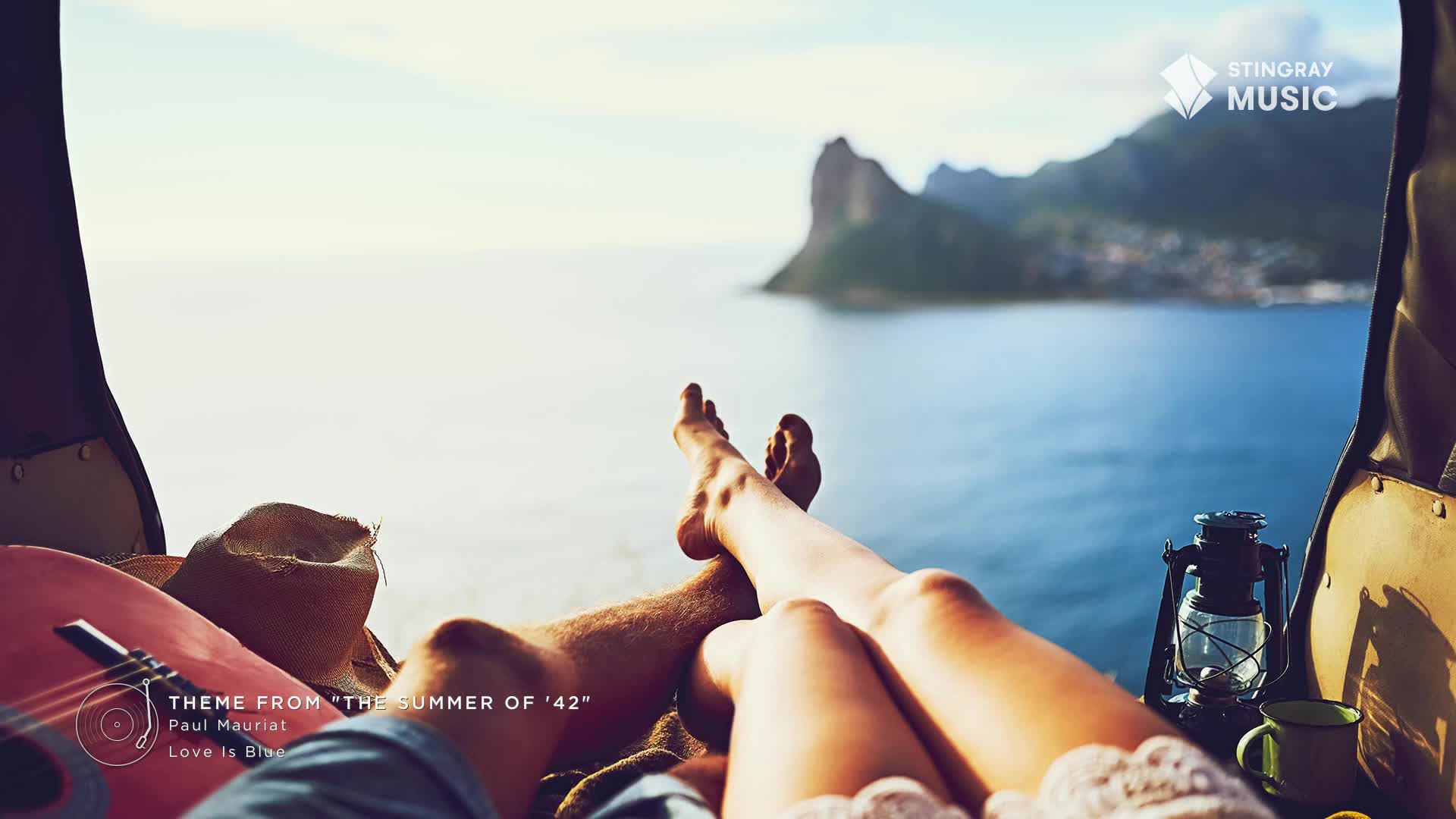 A couple relaxes, legs crossed, with a guitar and a lantern nearby, overlooking the ocean from what looks like a car trunk. The music of Stingray Easy Listening from Canada plays in the background.
