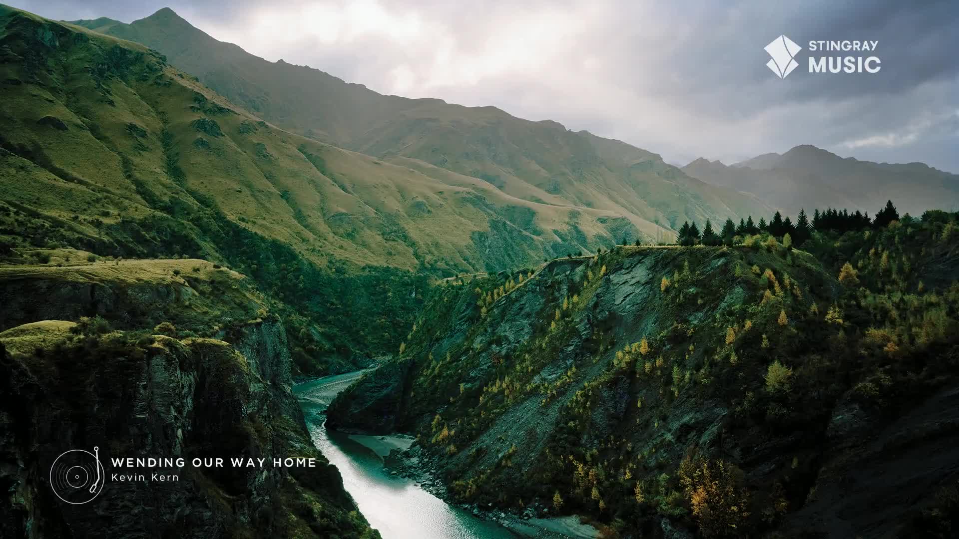A river carves its way through the deep valley, winding between steep, green mountains. The sky is overcast, but sunlight still illuminates the landscape, making the scene feel peaceful, like something you might hear on Stingray Easy Listening from Canada.
