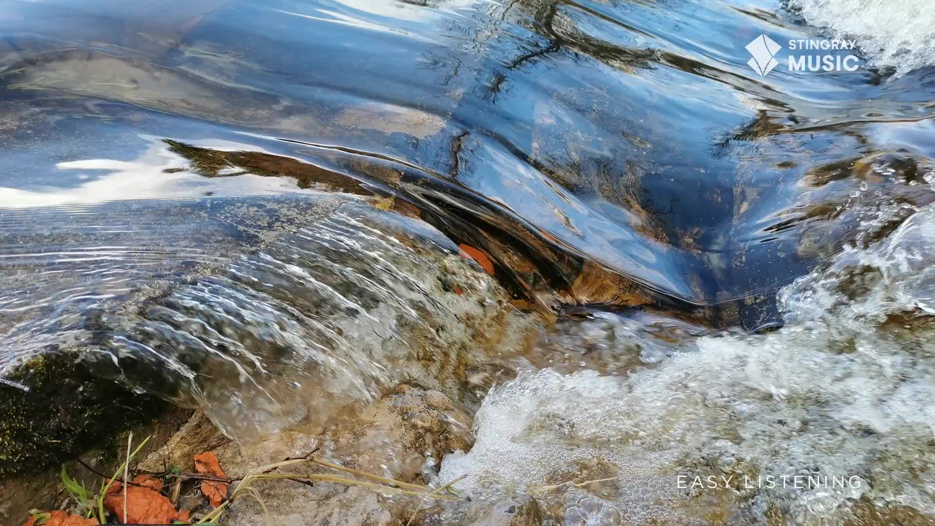 Water rushes over a small rock formation, creating a cascade of white foam. The surface reflects the sky, and the sound is soothing, like the music on Stingray Easy Listening.

