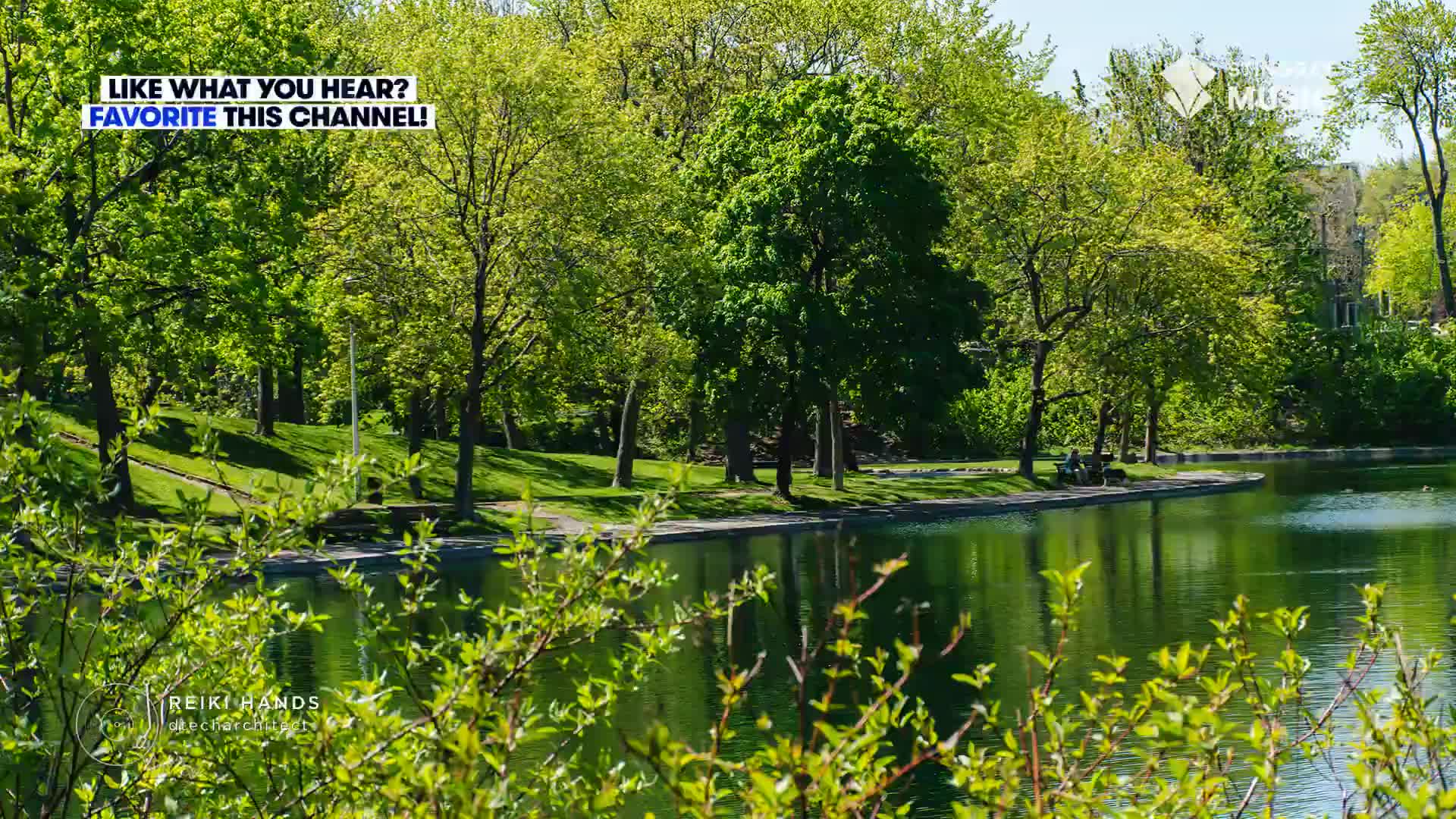 The lake's surface reflects the bright green trees lining the shore. Two people sit on a bench near the water's edge, enjoying the sunny day.
