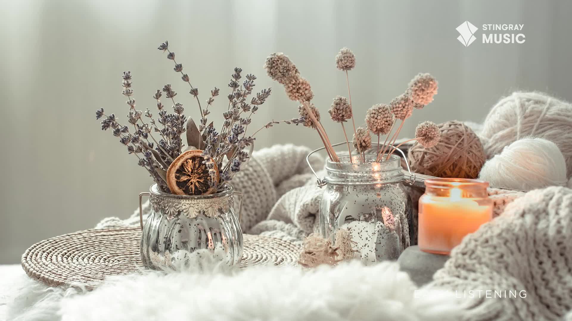Dried lavender and a slice of orange sit in a small, ornate glass vase. A candle's warm glow illuminates the scene, casting shadows over the soft textures of yarn and a woven mat.
