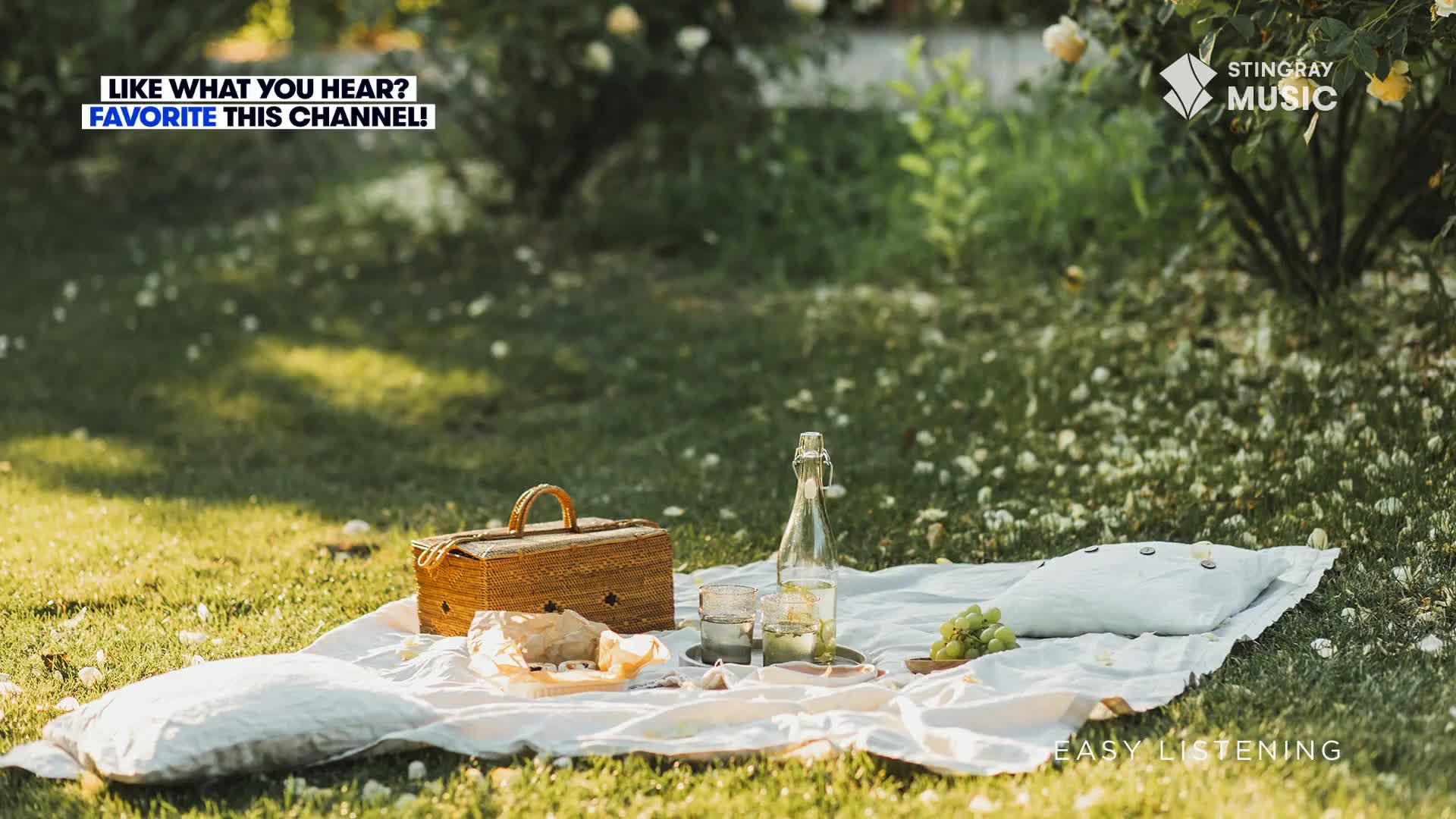 A picnic basket sits open on a white blanket, alongside a bottle of water and a few glasses. The soft grass and sunlight suggest a peaceful afternoon, perfect for listening to some easy listening music from Canada.
