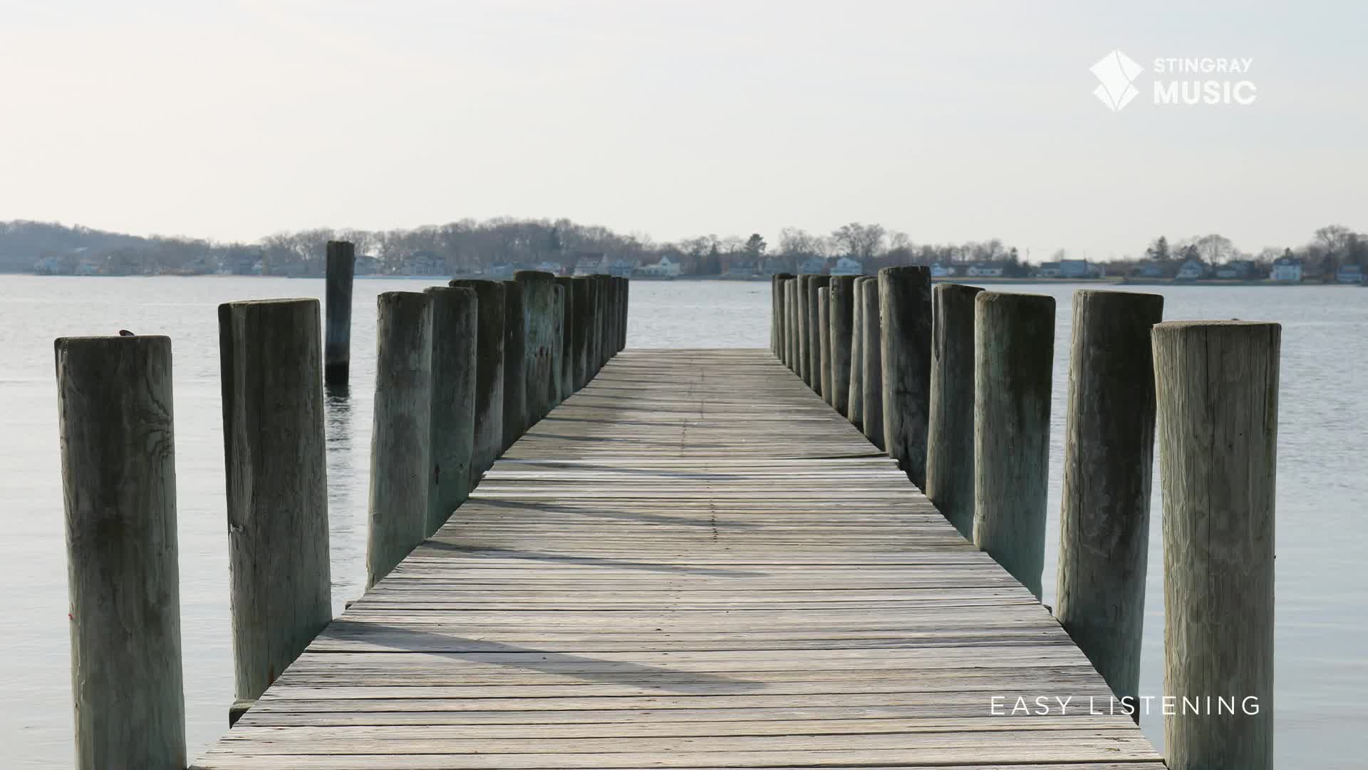 The wooden planks of a pier stretch out over the water, framed by weathered posts. Calm water reflects the sky, giving the scene a peaceful, Stingray Easy Listening vibe, like something you'd hear from Canada.
