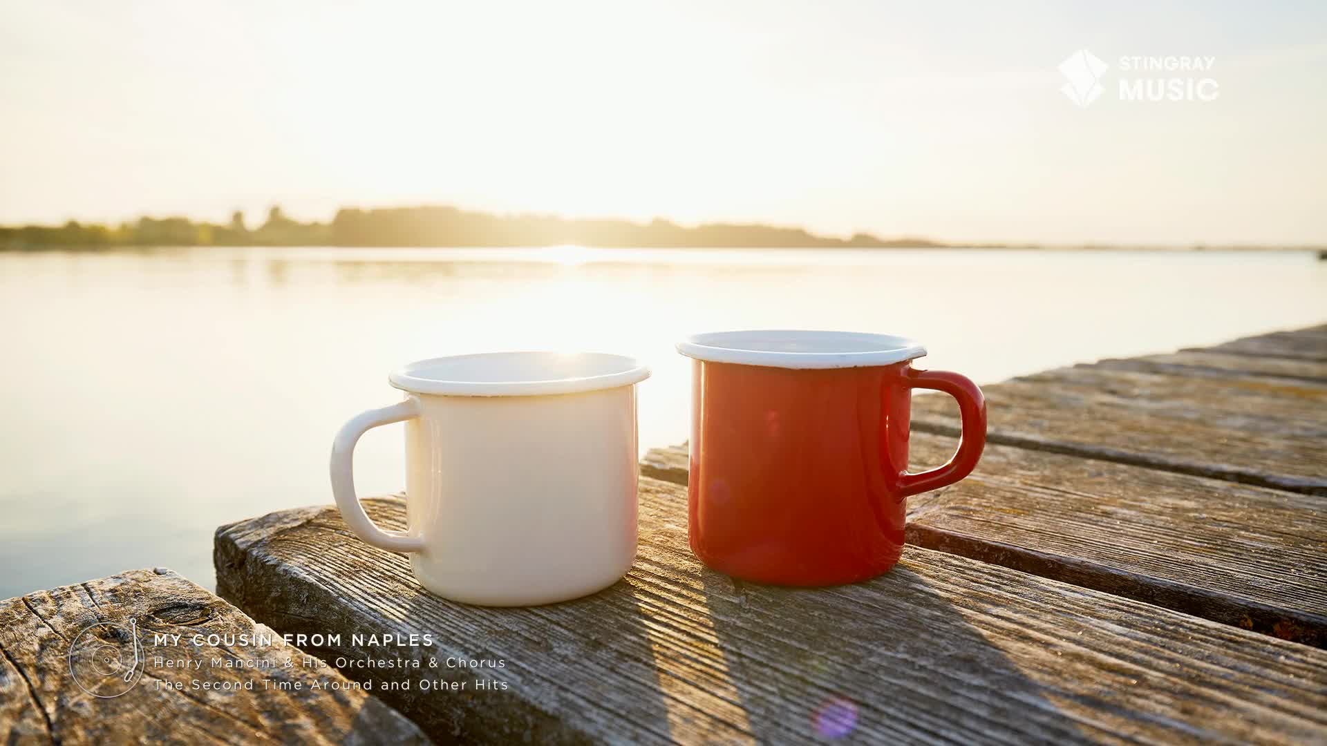 Two enamel mugs, one cream and one red, sit side-by-side on a weathered wooden dock, facing a calm Canadian lake. The Stingray Music logo sits in the corner, and the album title "My Cousin From Naples" is visible at the bottom, suggesting a moment of peaceful listening.
