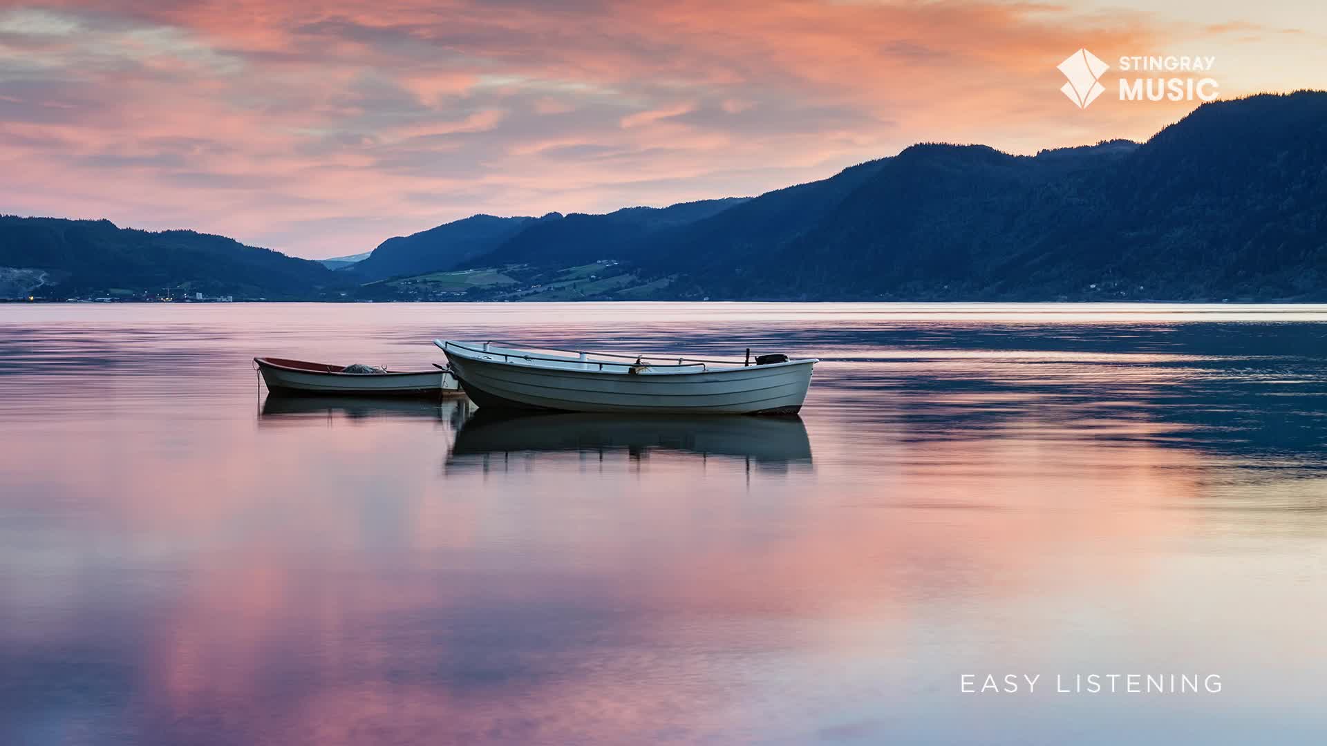 Two small boats gently bob on the glassy water, reflecting the soft pink and purple hues of the sky. The mountains in the distance are a deep blue, a perfect backdrop for the Canadian Stingray Easy Listening scene.

