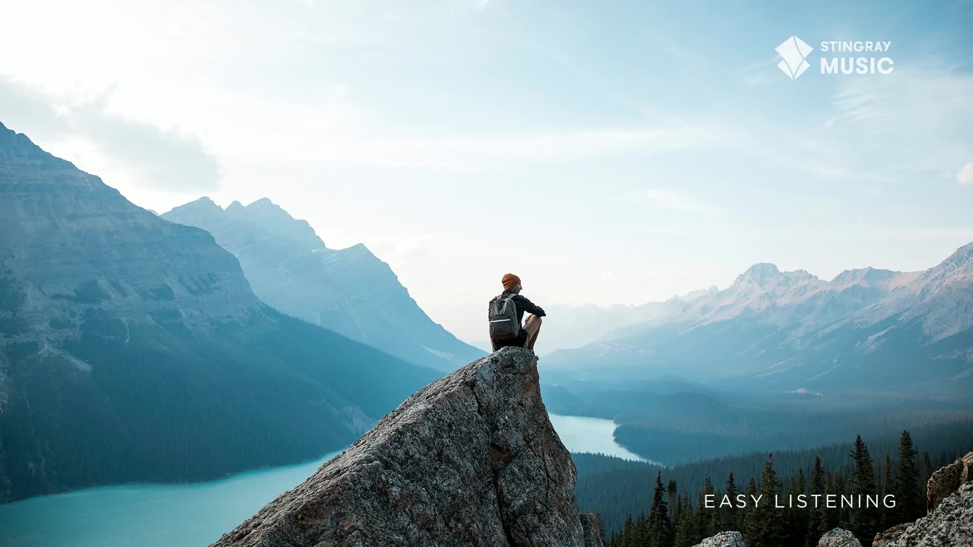 A lone figure sits perched on a rocky outcrop, gazing out at the vast Canadian landscape. The scene is serene, with the water below and the mountains in the distance, evoking a sense of calm that aligns with the Stingray Easy Listening vibe.
