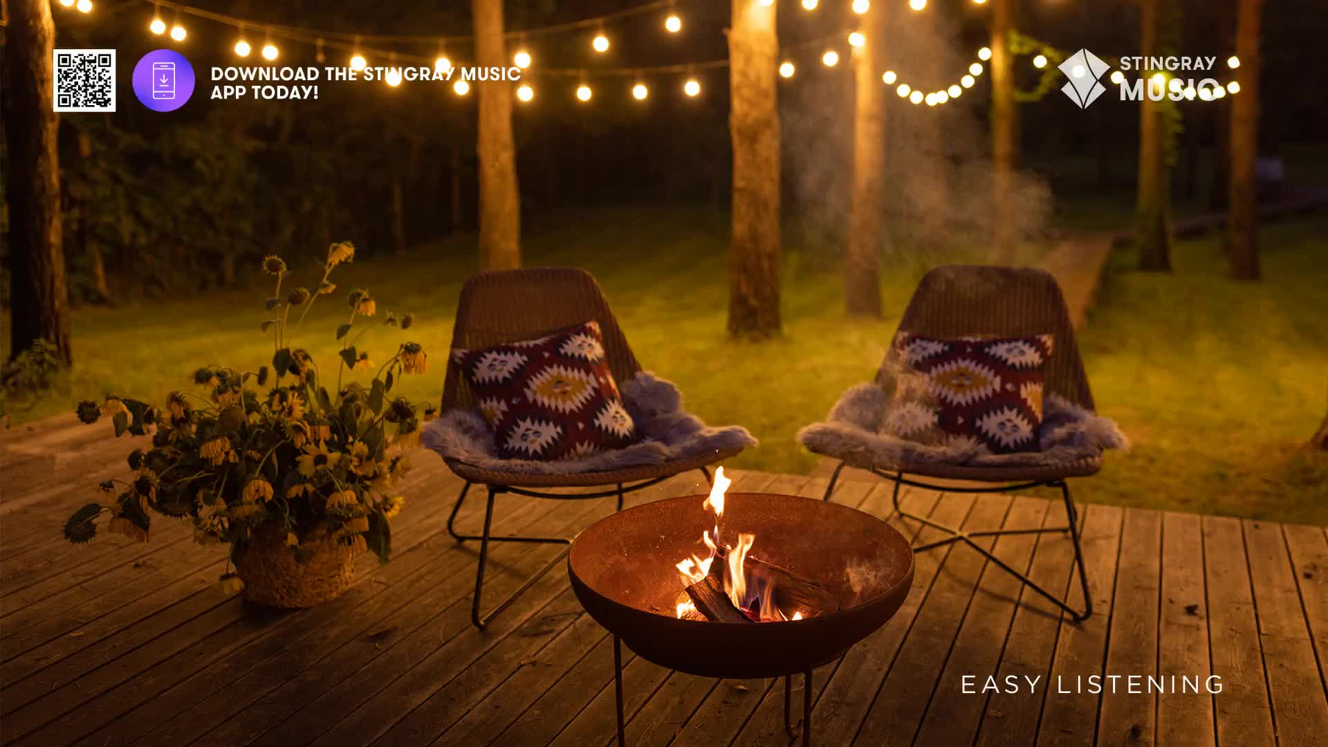 The fire crackles in a metal bowl, sending wisps of smoke up into the night air. Two chairs sit ready, each with a patterned pillow and a soft, furry throw, under the glow of string lights.
