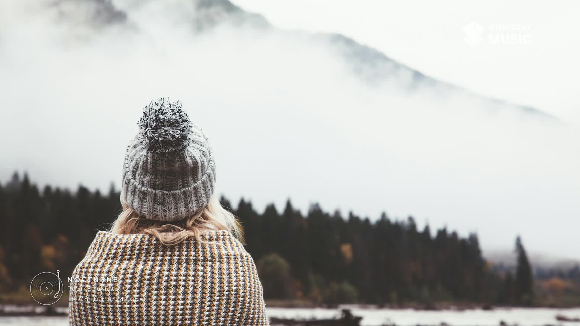 The fog hangs thick over the mountains, obscuring the peaks. A woman in a knit cap and patterned wrap gazes out at the misty landscape, the scene evoking the calming sounds of Stingray Easy Listening.
