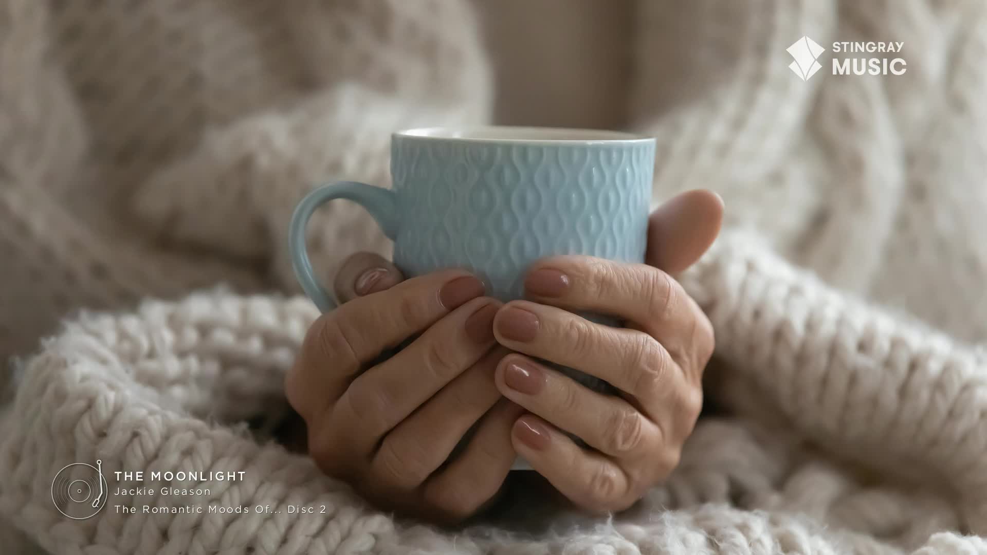 Hands, with polished nails, cradle a light blue mug, nestled in a thick, cream-colored sweater. The Stingray Music logo is visible, perhaps the soundtrack to a cozy moment.
