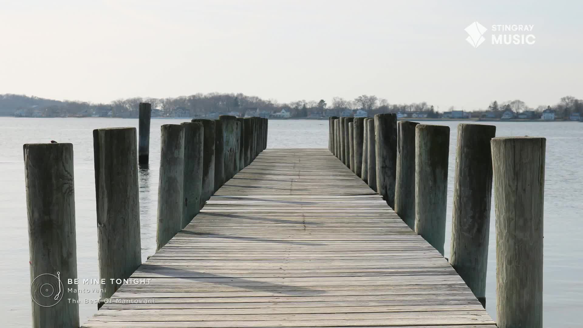 A weathered wooden pier stretches into calm water, the pilings casting long shadows. The Stingray Music logo sits in the top right corner, a reminder of the easy listening tunes that would be perfect for this scene.
