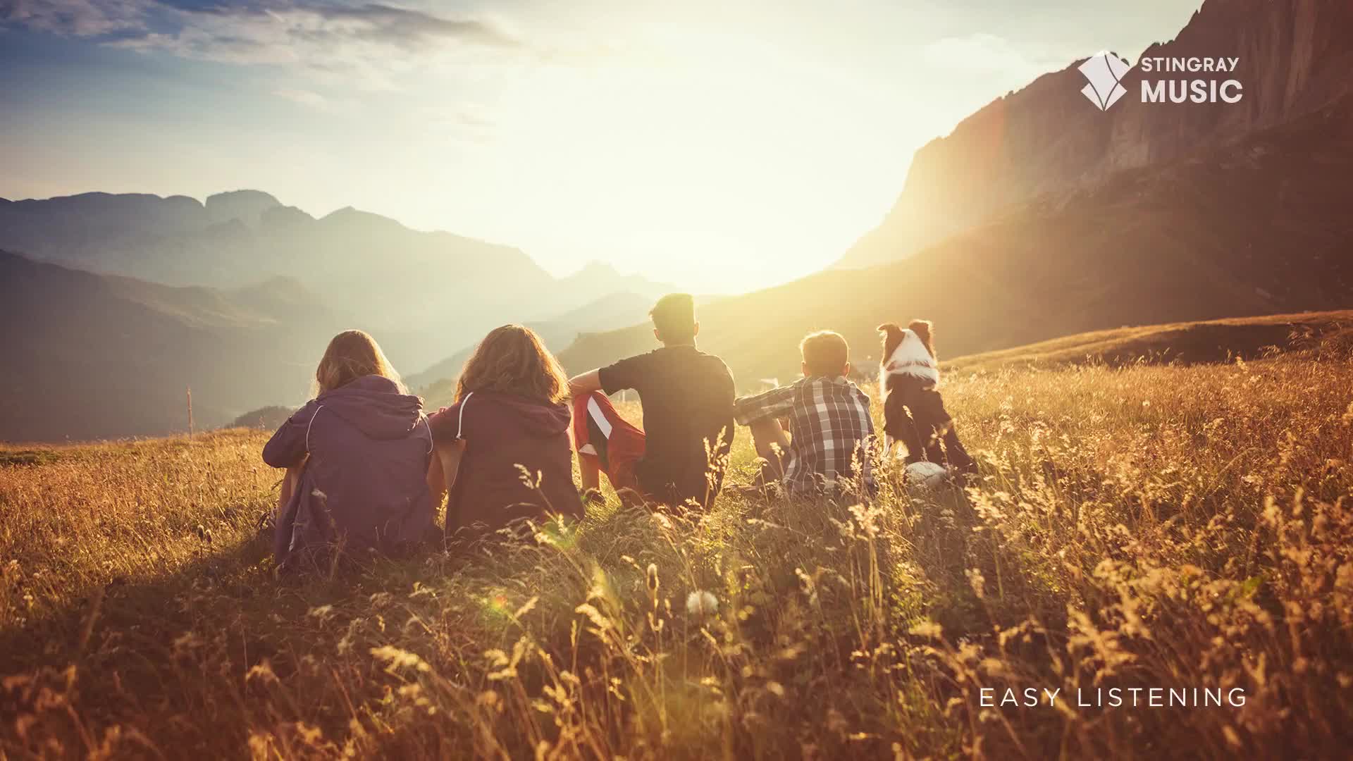 A group of people, plus a dog, are sitting in a field, backs to the camera, gazing at the mountains. The sun is setting behind the peaks, casting a warm glow over the scene, which feels like a perfect fit for Stingray Easy Listening.
