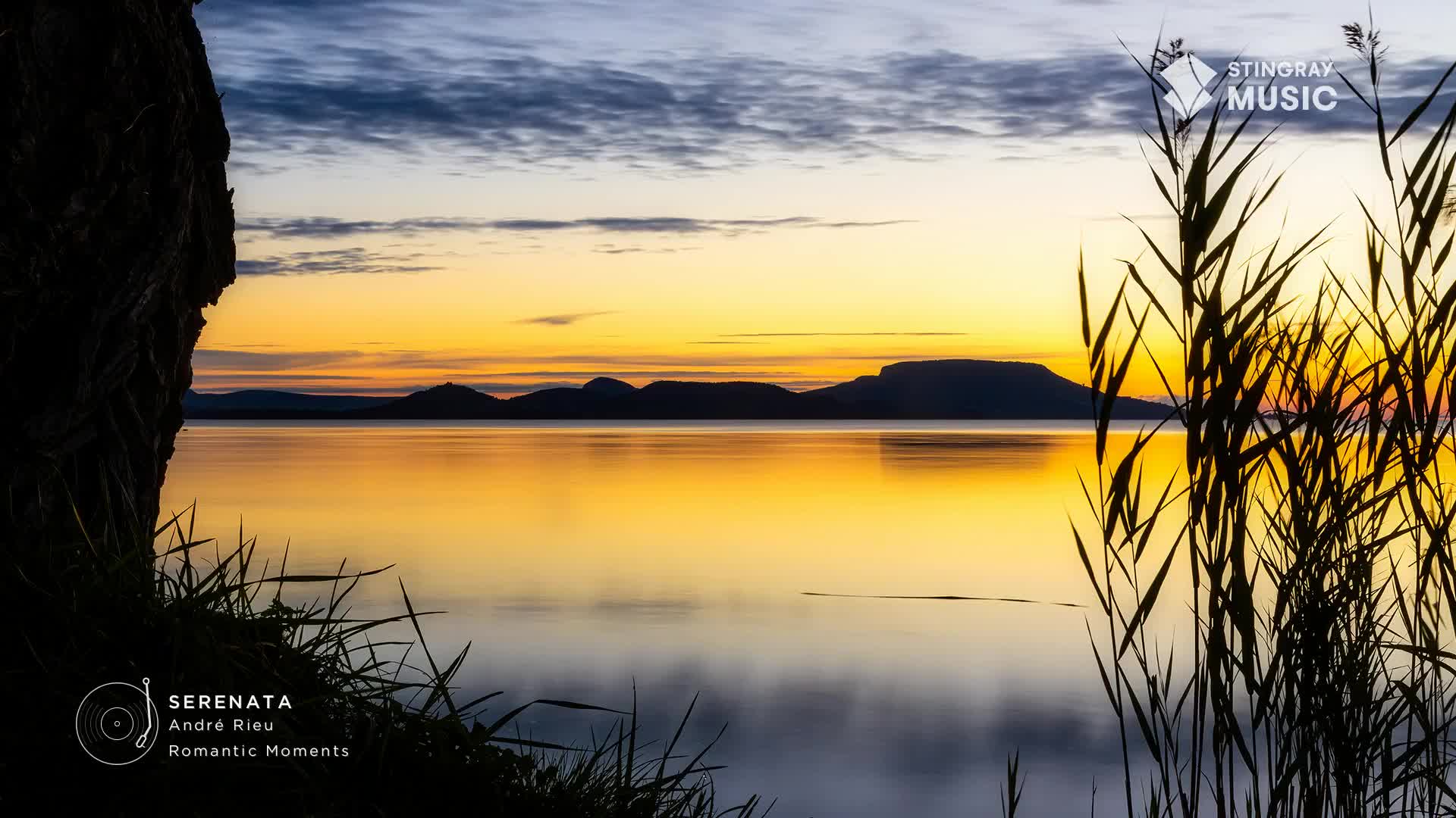 The water reflects the fiery sunset, with the sky and mountains mirrored in its stillness. Reeds sway gently in the foreground, as if accompanying the easy listening music from Stingray in Canada.

