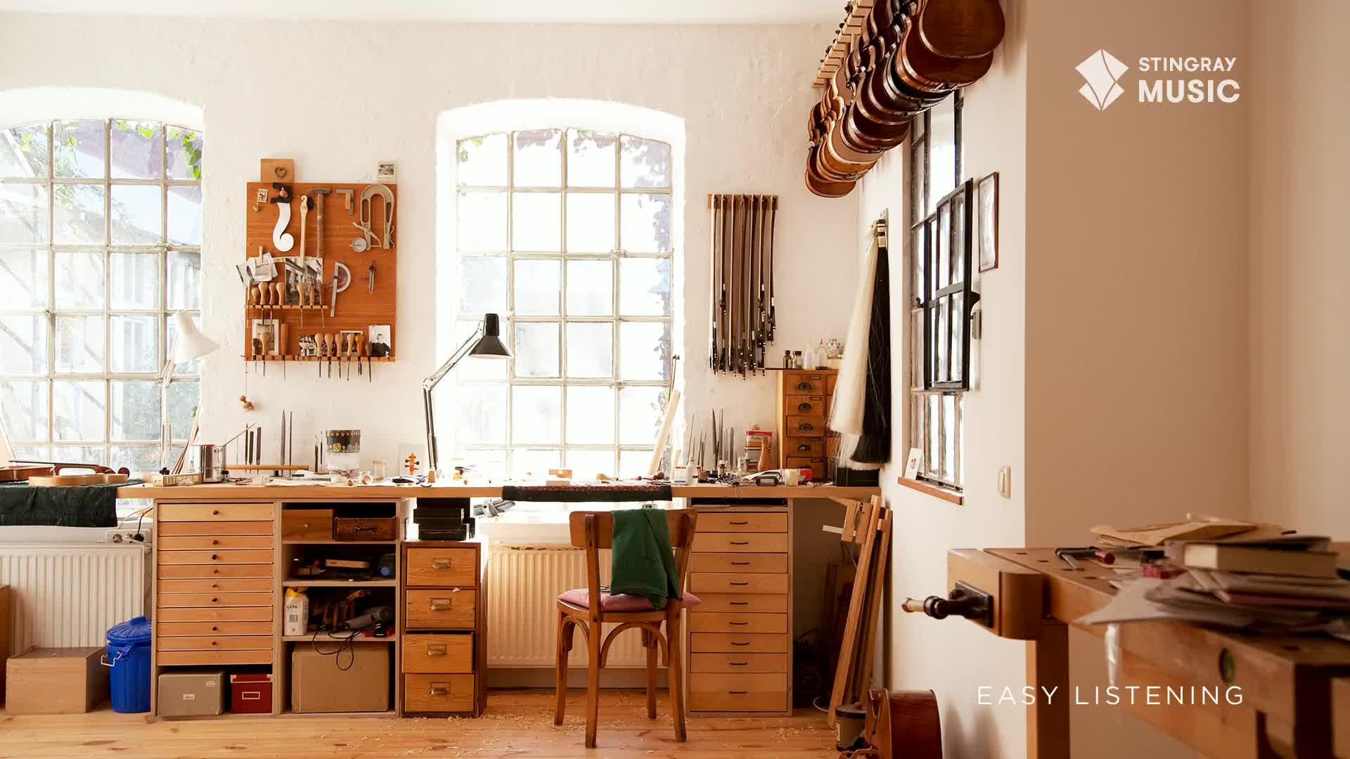 Sunlight streams through the large windows, illuminating a violin maker's workshop, presumably somewhere in Canada, as Stingray Easy Listening plays softly in the background.  A collection of violins hangs above the window, and the workbench is filled with tools and materials.

