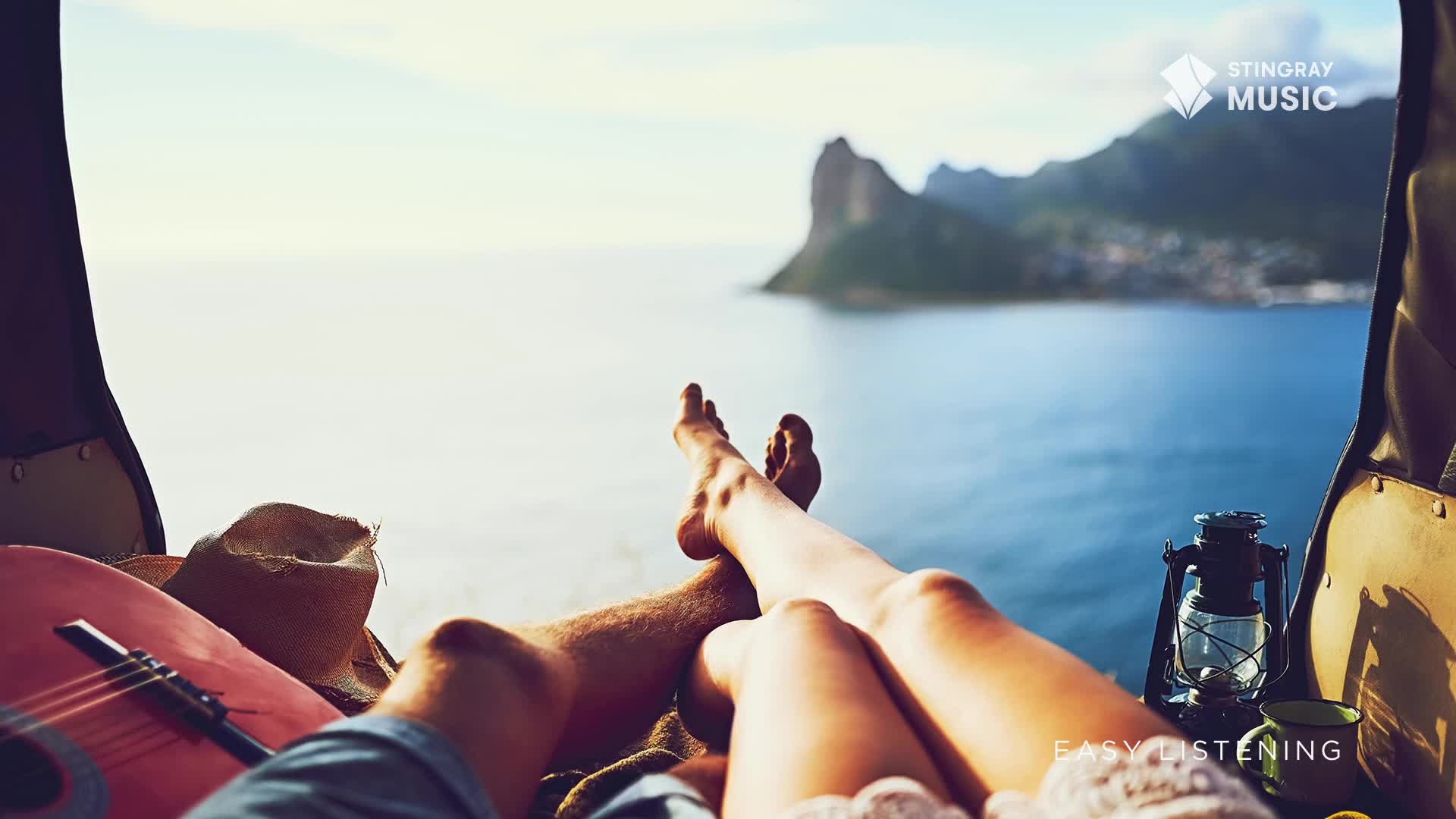 Bare legs are crossed, feet pointing towards the ocean, framed by the open back of a vehicle. The scene, perfect for Stingray Easy Listening, unfolds against a backdrop of water and mountains.
