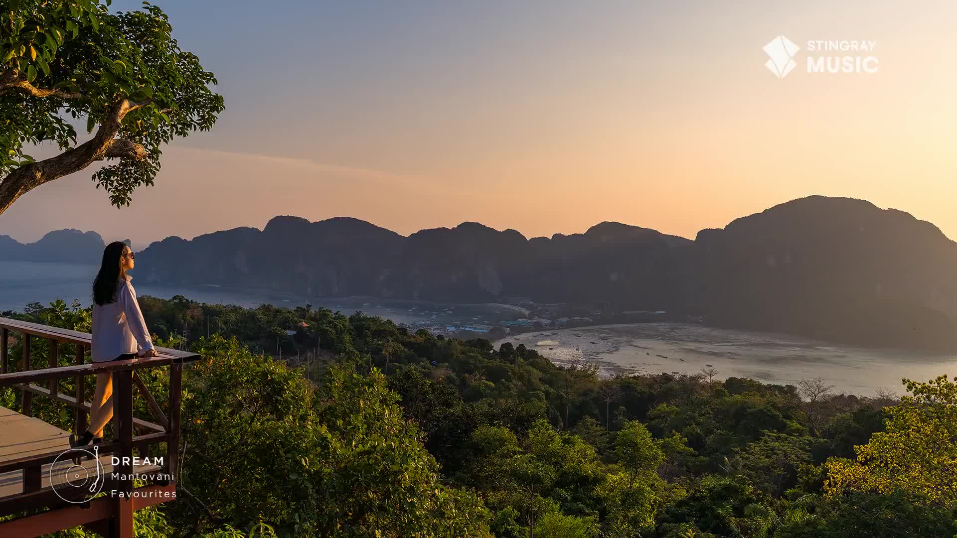 A woman stands on a wooden deck, gazing at a bay surrounded by mountains. The sky is a soft gradient of orange and blue, a perfect backdrop for the scene.
