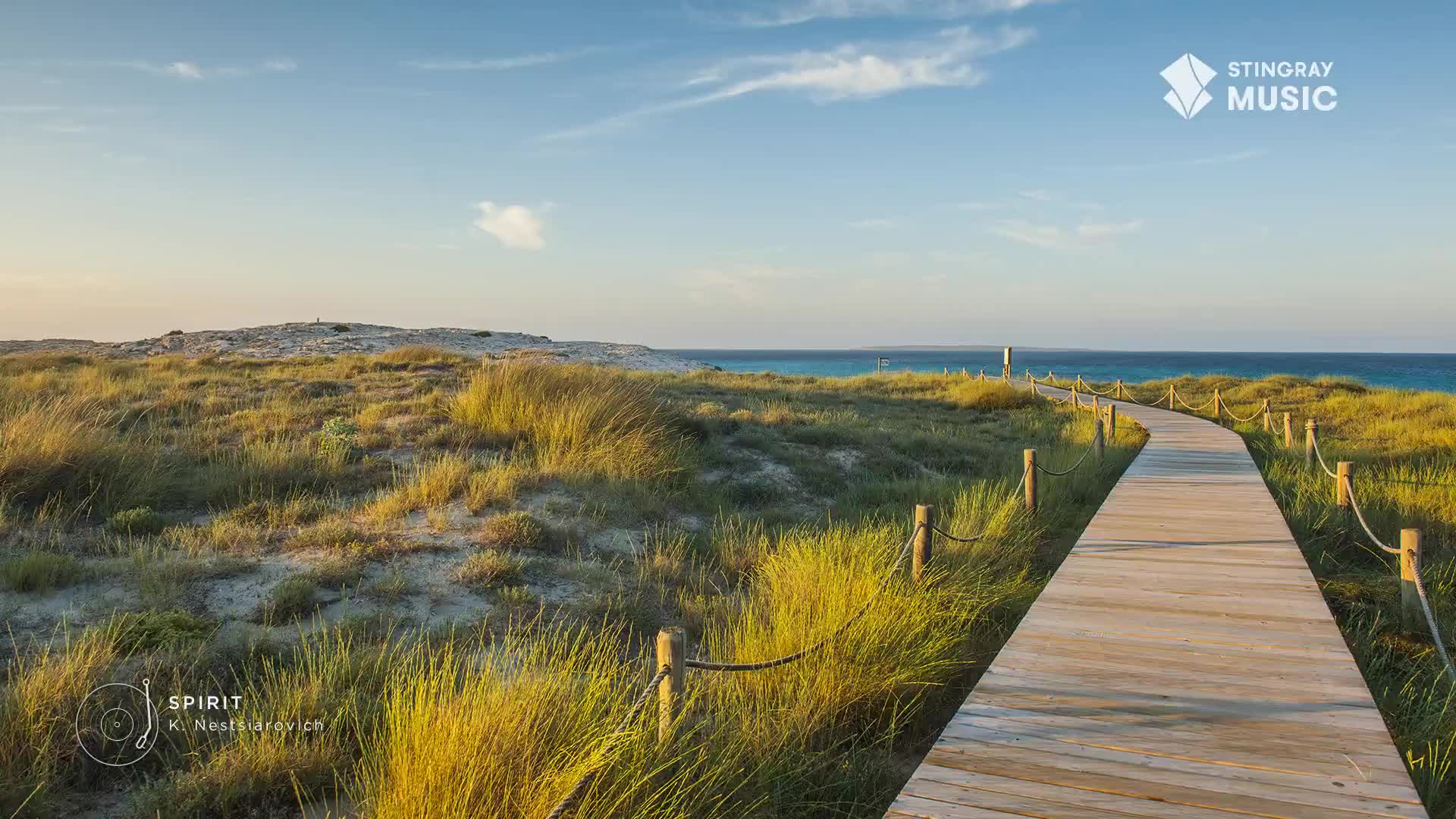 A wooden boardwalk leads toward the ocean, flanked by grassy dunes. The sky is a clear blue, and the scene feels like something you'd hear on Stingray Easy Listening from Canada.
