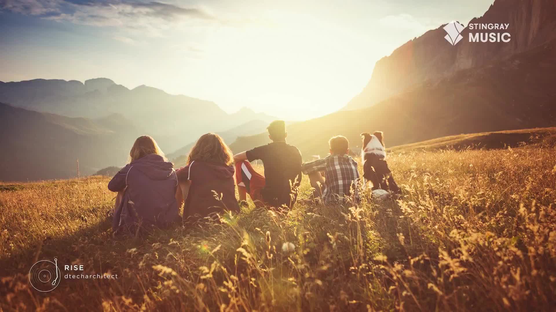 A group of people, backs to the camera, sit in a field of tall grass, gazing at mountains bathed in sunlight. A dog sits patiently beside them, enjoying the view.
