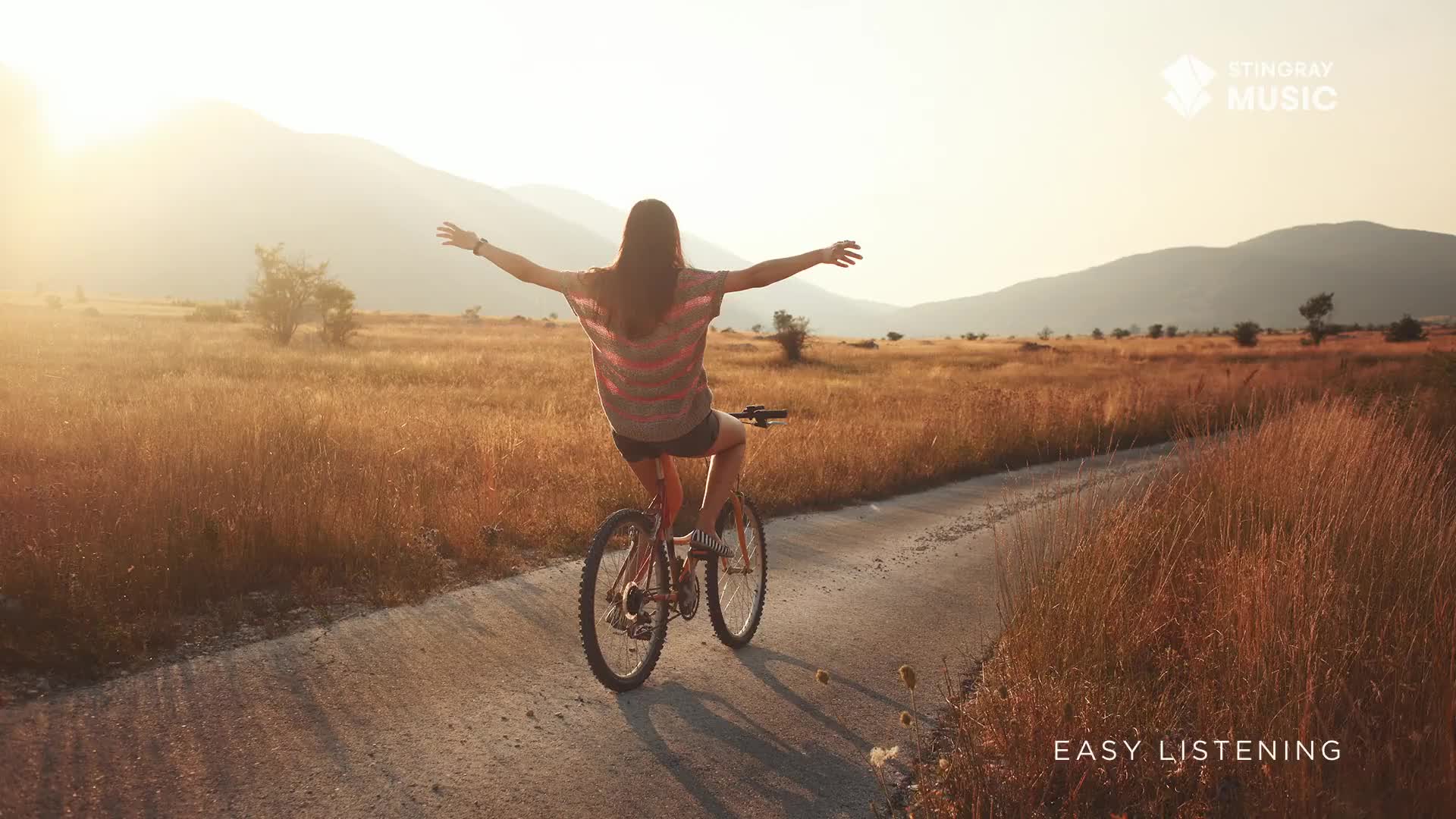 A woman rides a bicycle down a sunlit path, arms outstretched. The landscape is a field of tall, dry grass, and the scene evokes the gentle sounds of Stingray Easy Listening.
A woman rides a bicycle down a sunlit path, arms outstretched. The landscape is a field of tall, dry grass, and the scene evokes the gentle sounds of Stingray Easy Listening.