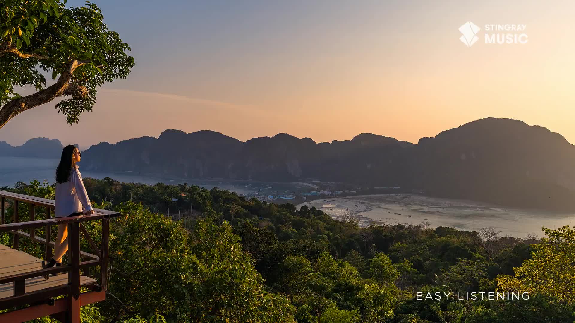 A woman stands on a wooden deck, gazing at a bay framed by dark mountains. The sky is a warm gradient of orange and yellow, promising a peaceful evening, as Stingray Easy Listening might suggest.
A woman stands on a wooden deck, gazing at a bay framed by dark mountains. The sky is a warm gradient of orange and yellow, promising a peaceful evening, as Stingray Easy Listening might suggest.